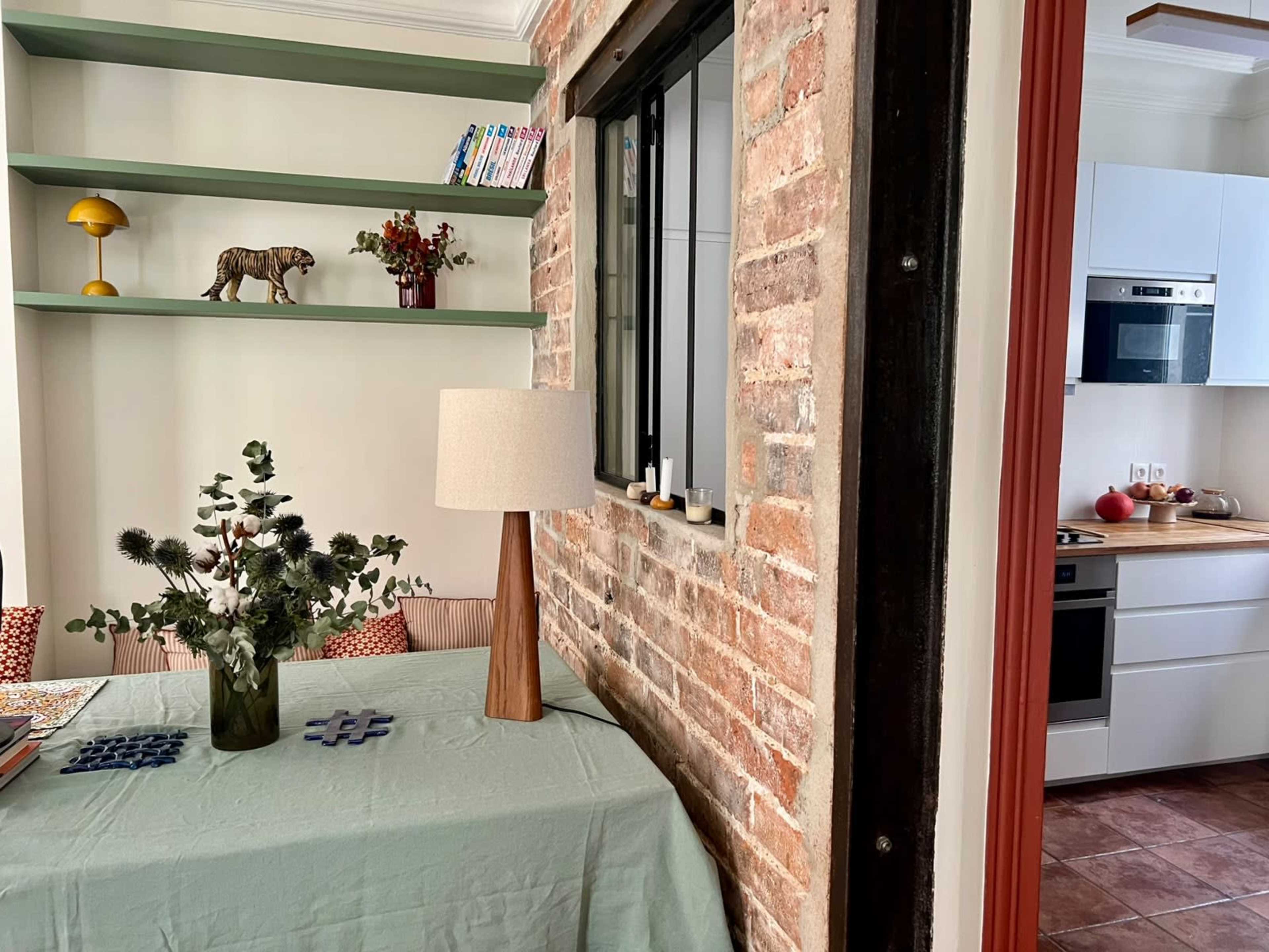 The image shows a cozy dining area featuring a wooden table with a green tablecloth, a decorative table lamp, a vase of greenery, and a wall with exposed brick that connects to a modern kitchen.