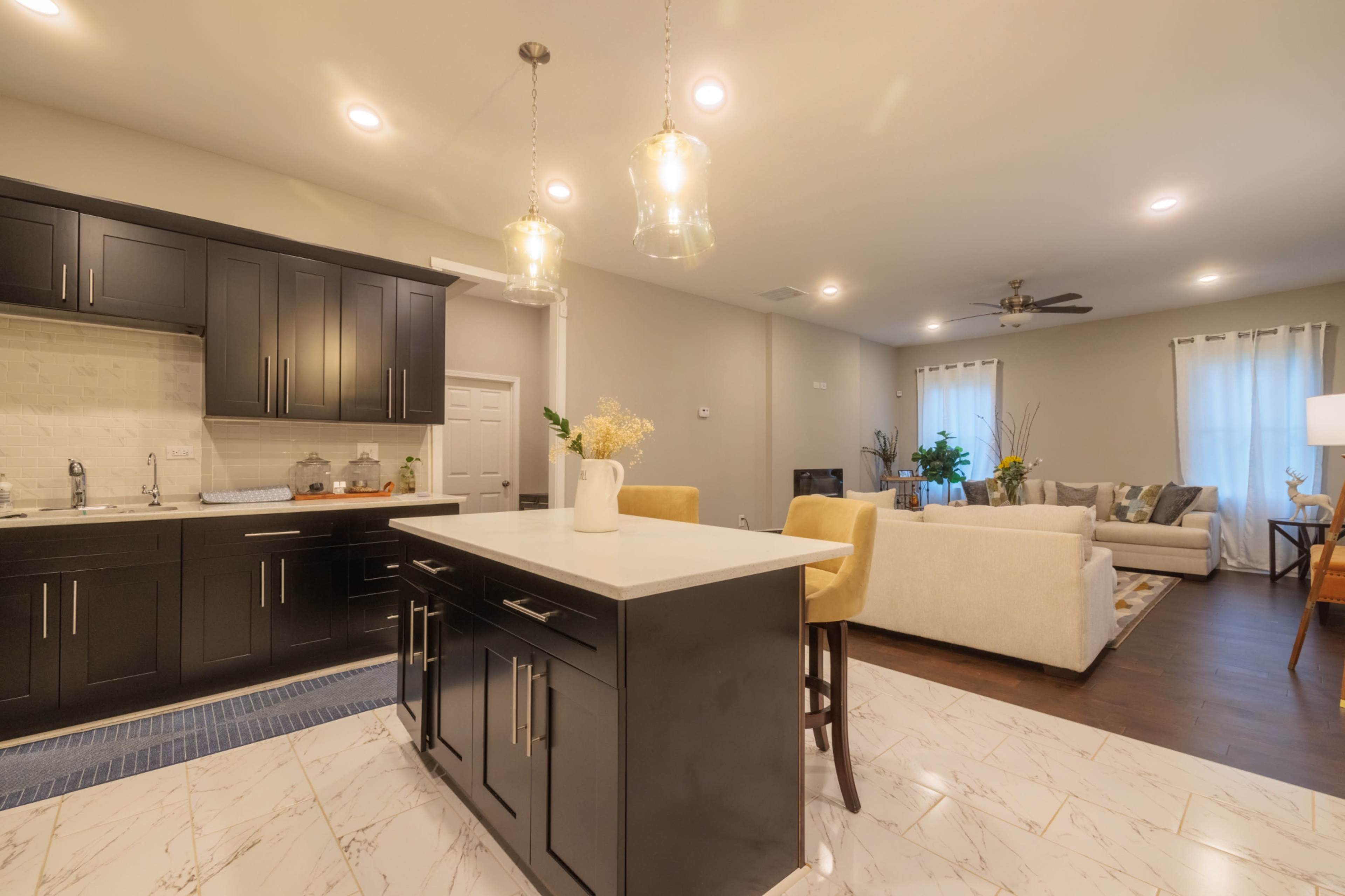 A modern kitchen with dark cabinetry, a central island with bar seating, and an adjoining living area furnished with a cream-colored sofa.
