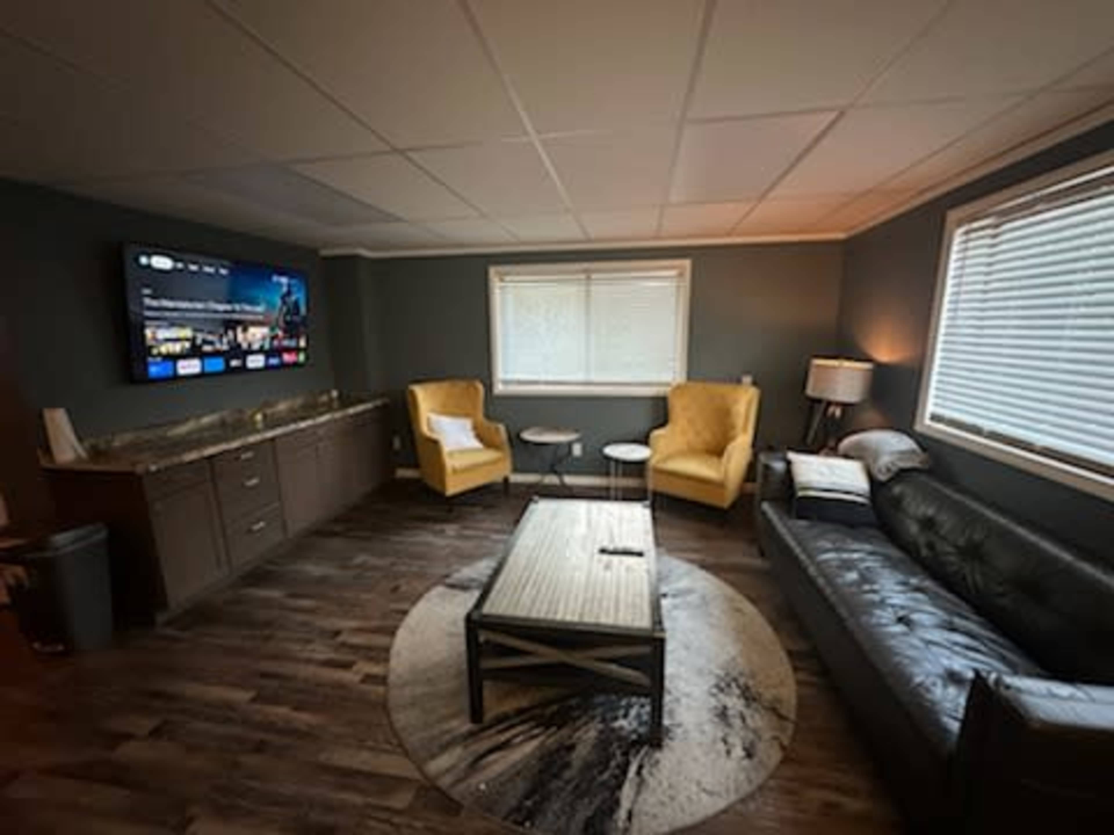 A modern living room with a black leather sofa, two yellow armchairs, a round rug, a coffee table, and a large wall-mounted TV.