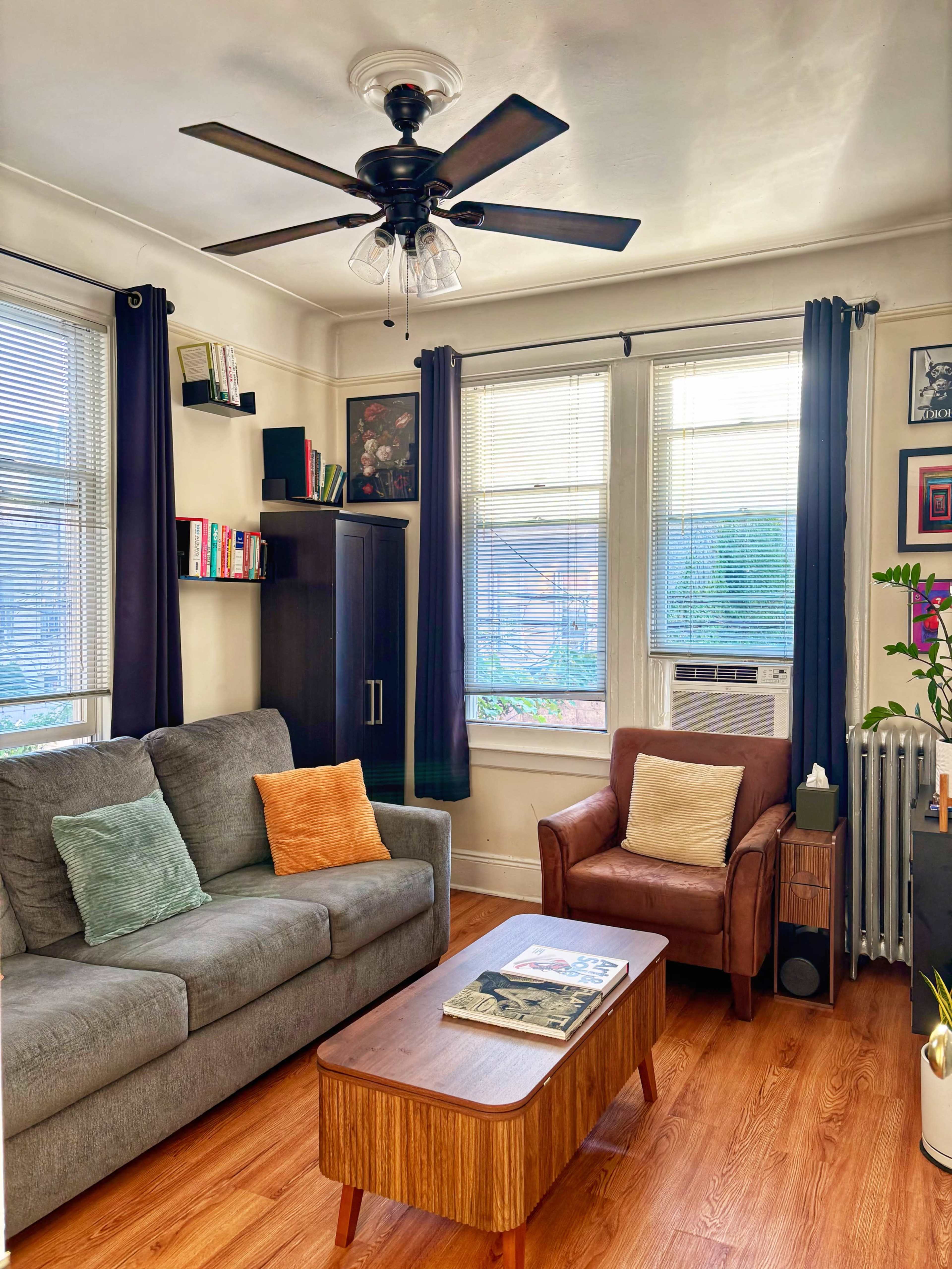 The image shows a cozy living room with a gray sofa, a brown armchair, a wooden coffee table, and large windows allowing natural light to enter the space.
