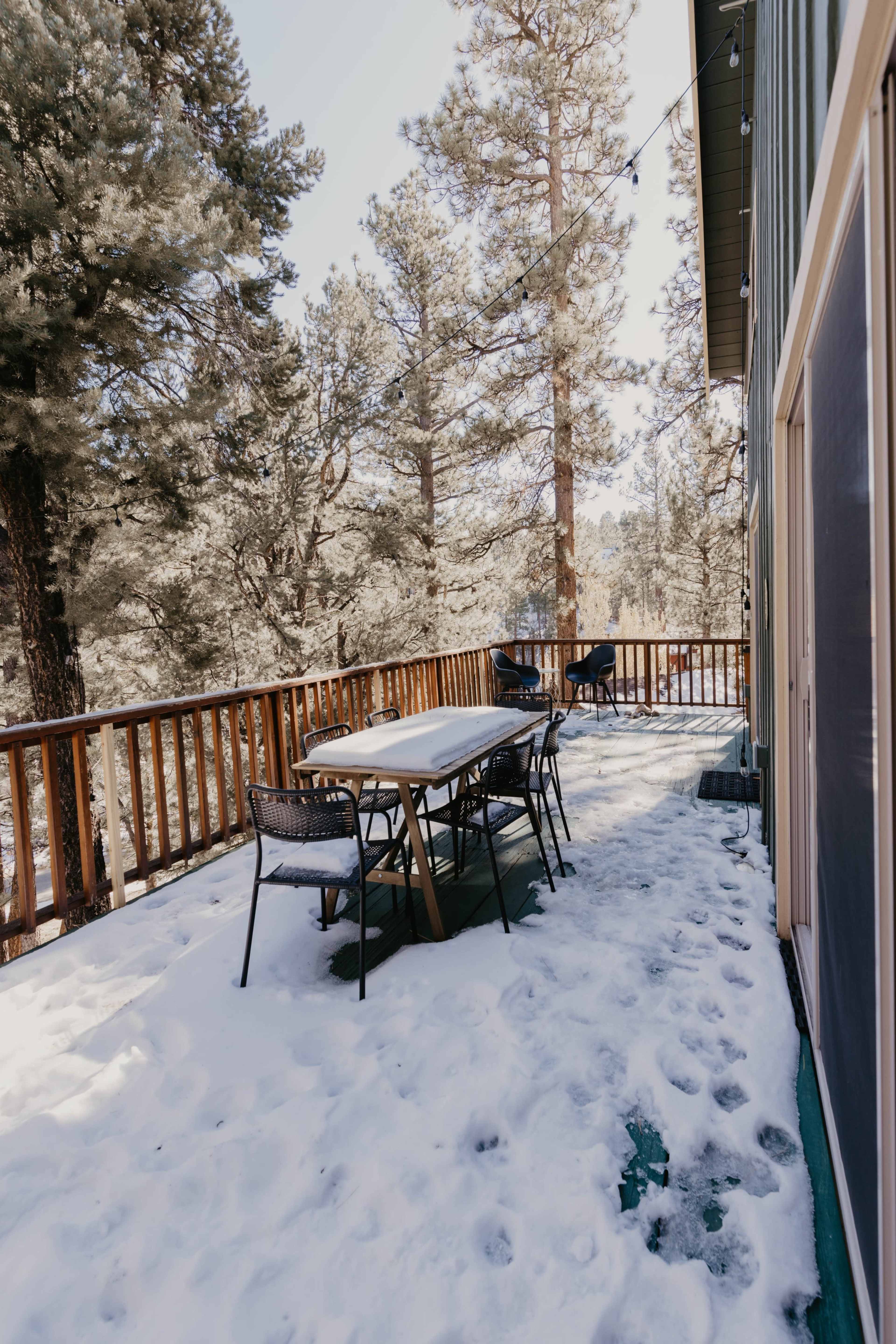 A wooden deck covered in snow features a table and chairs surrounded by tall trees.