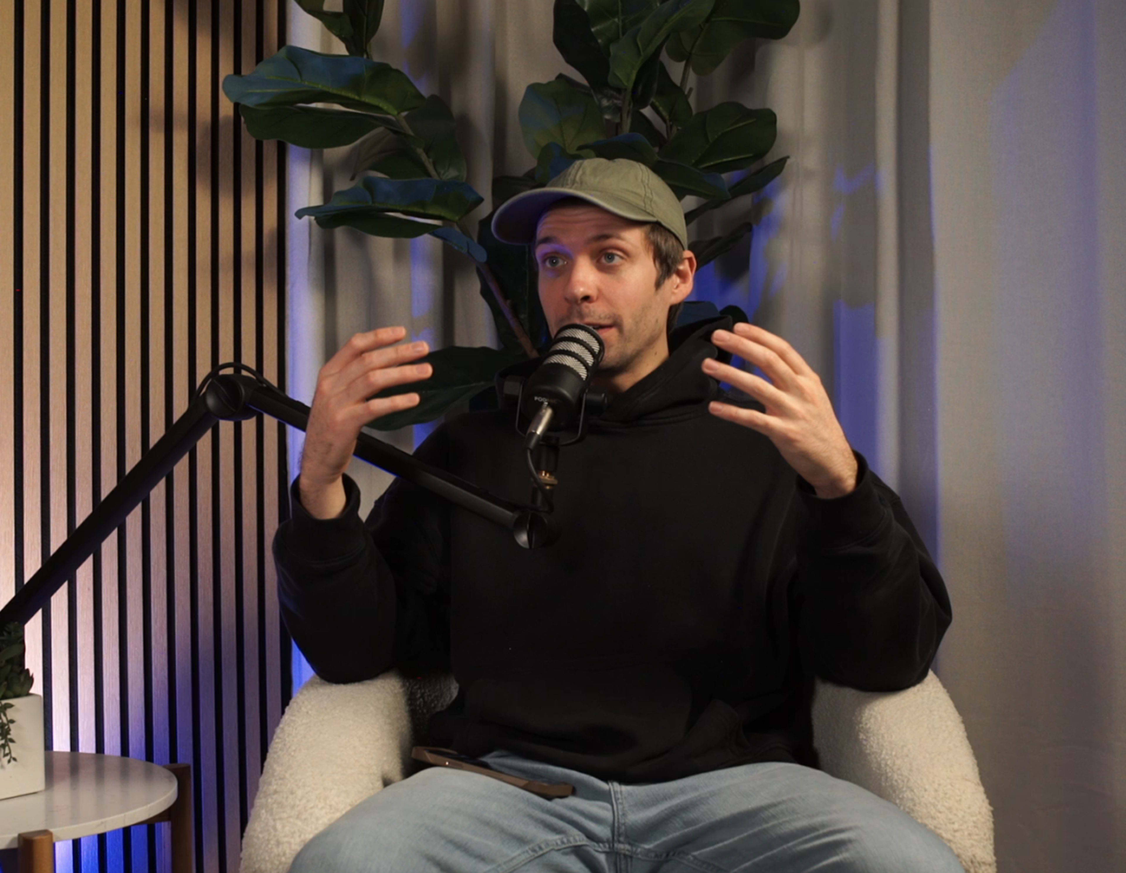 A person in a black hoodie is speaking into a microphone while sitting in a chair surrounded by indoor plants and wooden paneling.