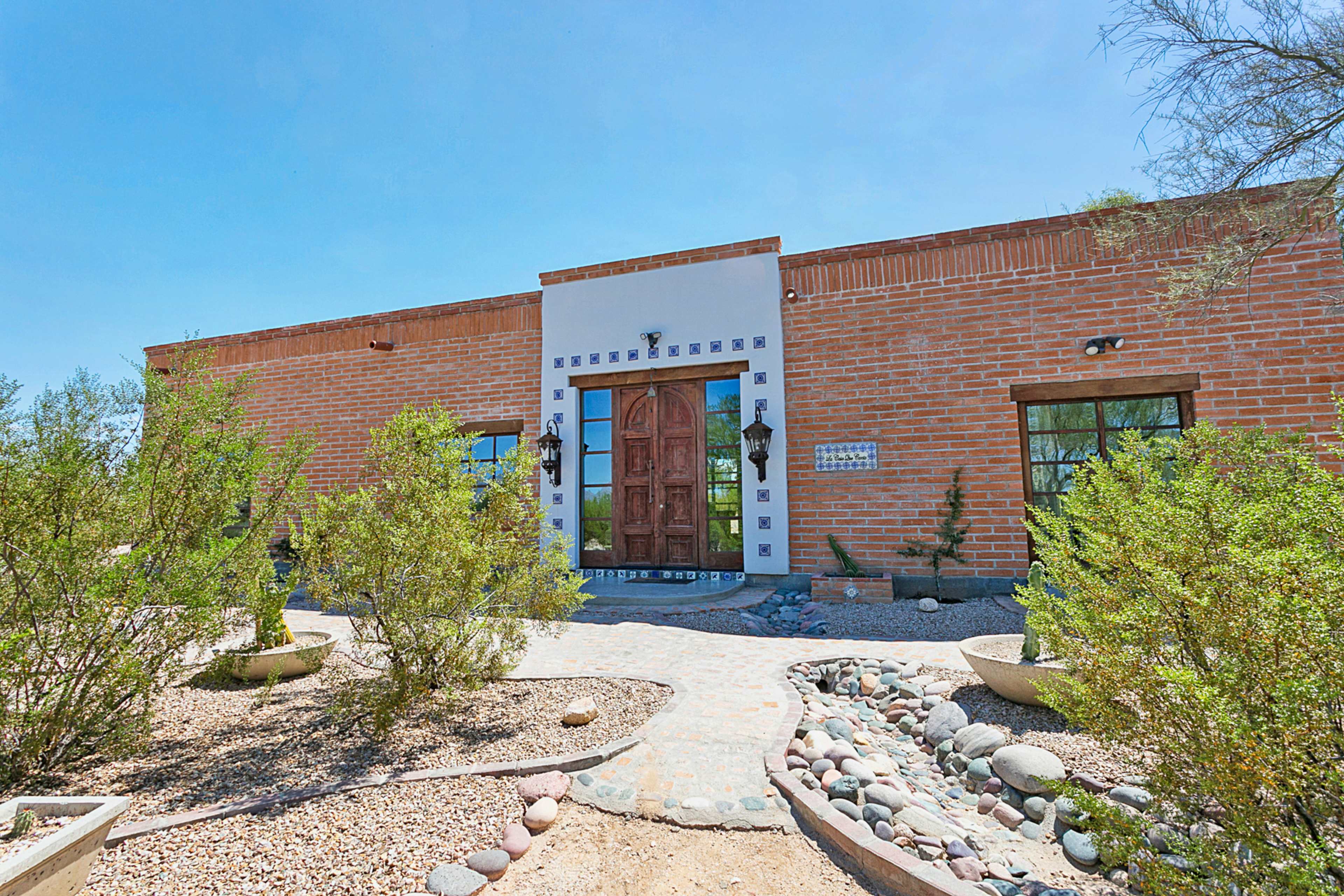 A single-story adobe building with a wooden door, surrounded by desert landscaping, including rocks and shrubs.