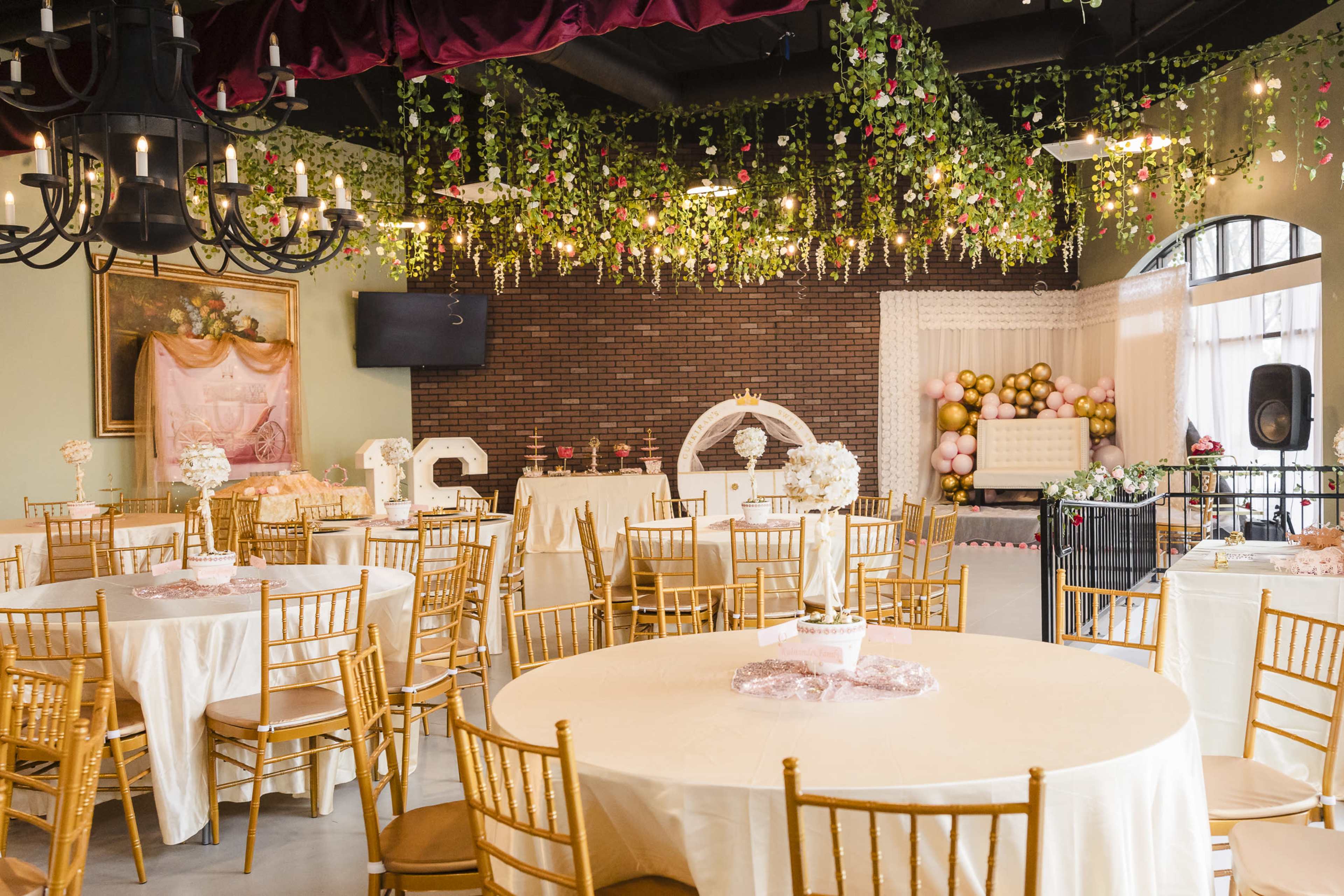 The image shows an elegantly decorated event space with gold chairs, round tables covered in white tablecloths, and floral arrangements hanging from the ceiling.