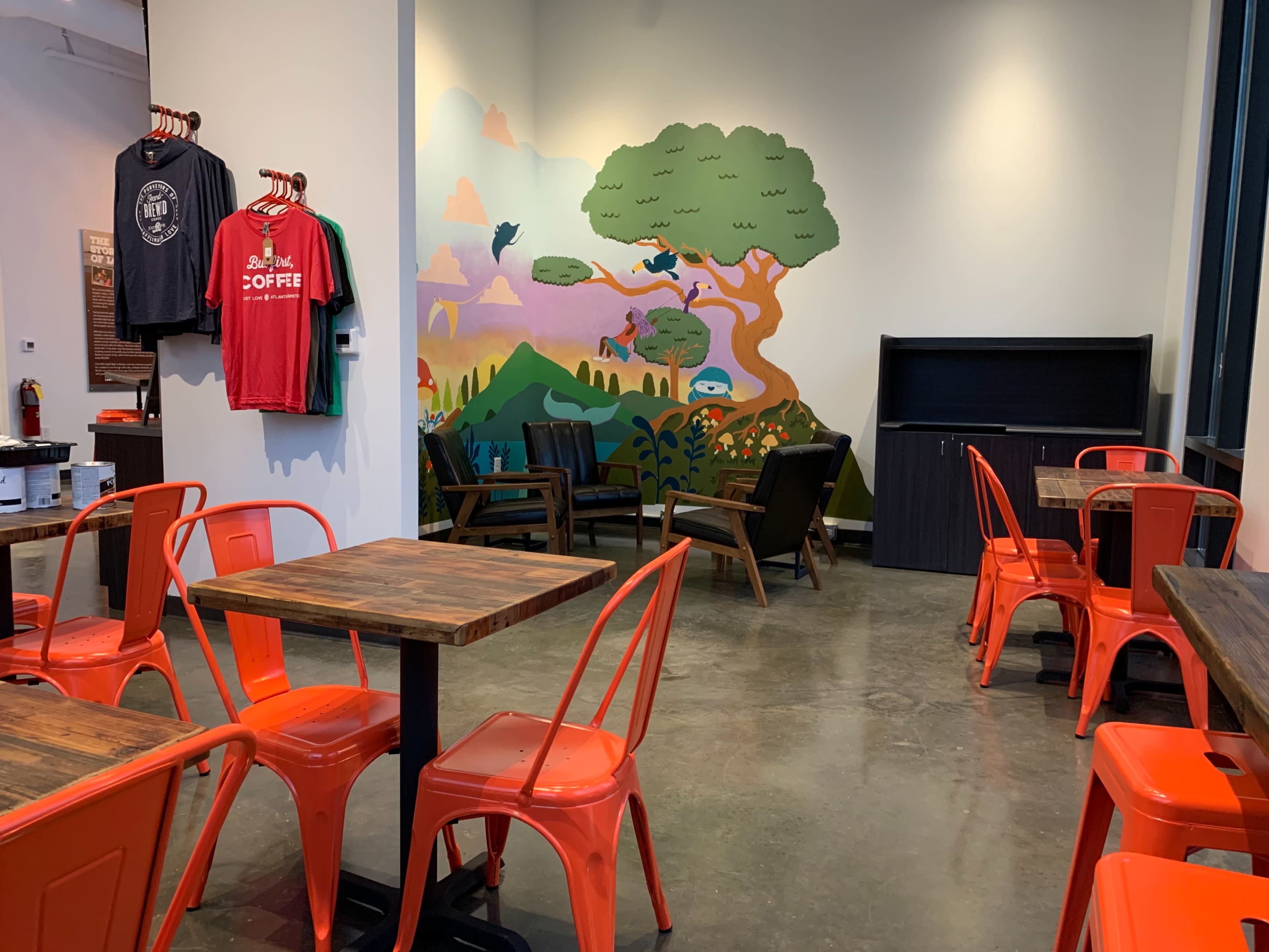 The image shows a café interior featuring orange metal chairs, wooden tables, and a colorful mural depicting a tree and outdoor scenes.
