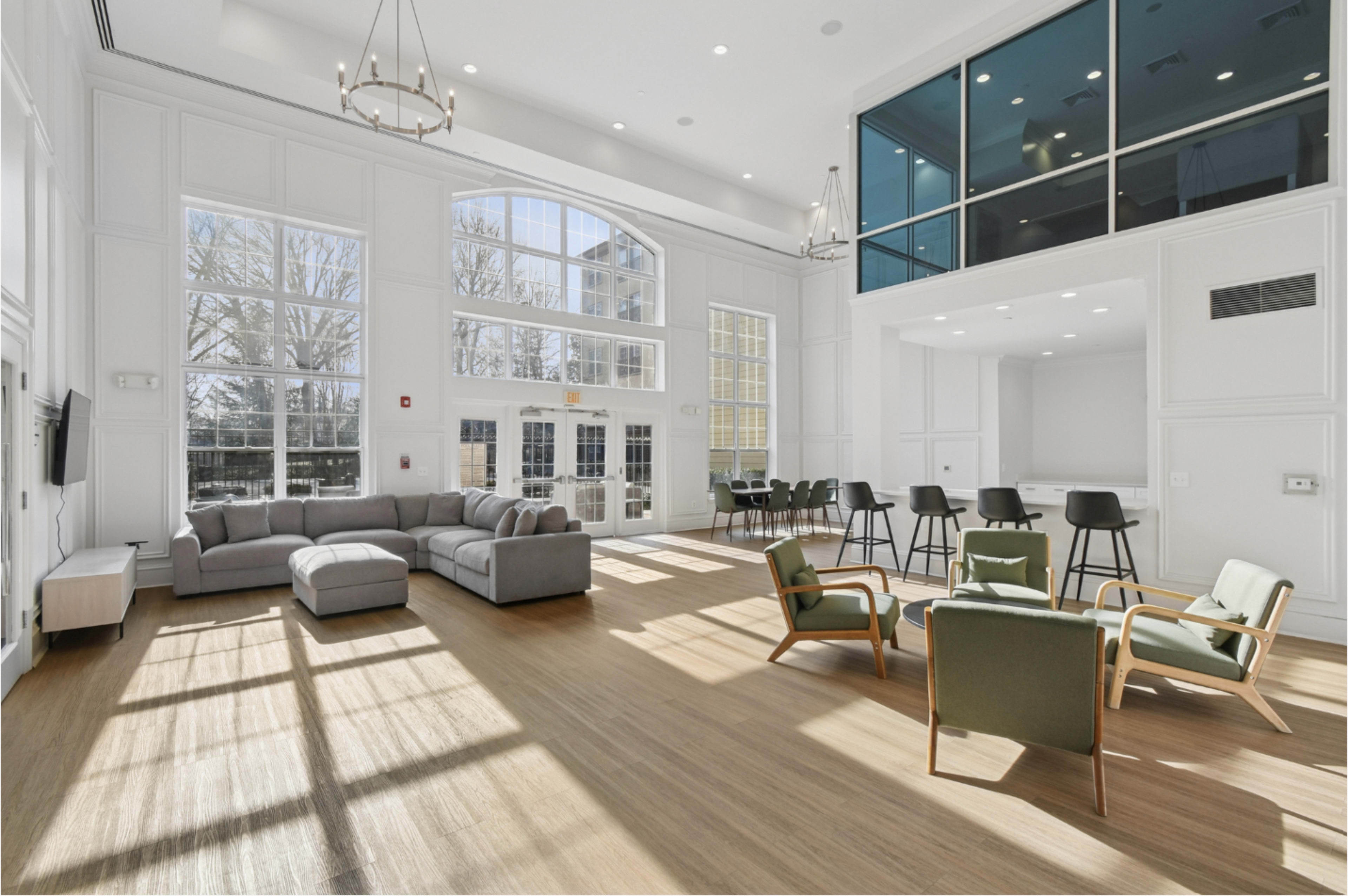 The image shows a spacious, well-lit common area with large windows, featuring a gray sectional sofa, green armchairs, and a dining area with barstools.