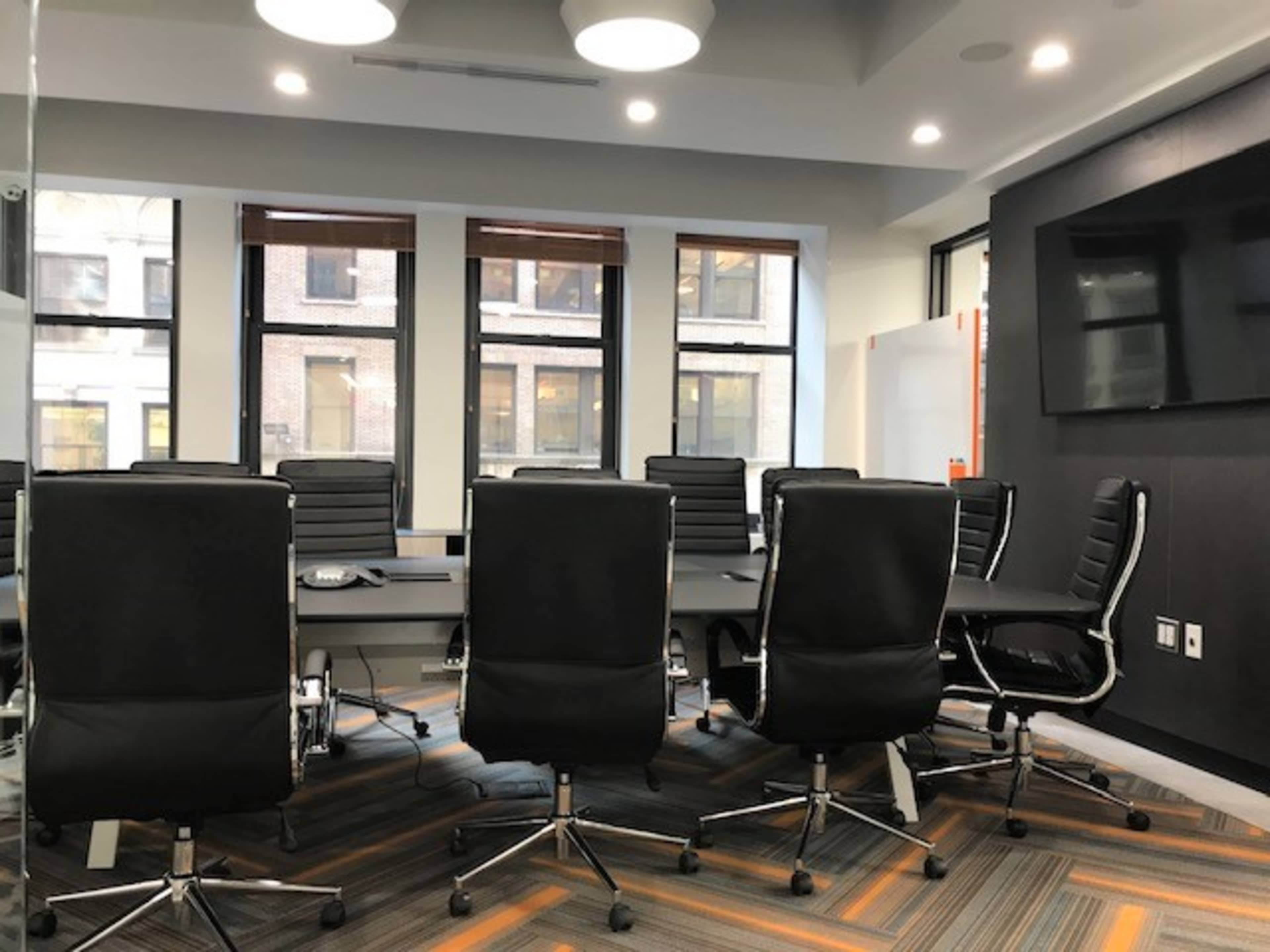 A modern conference room features a long table surrounded by black rolling chairs and large windows letting in natural light.