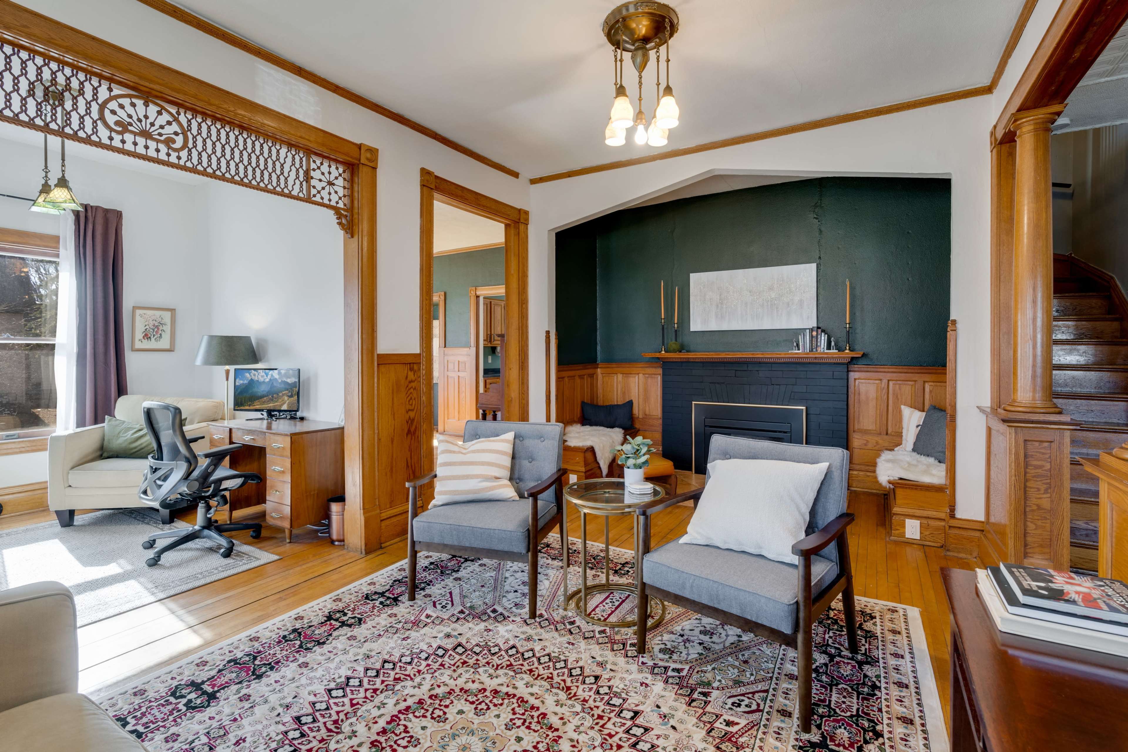 A cozy living room features two armchairs, a small side table, and a decorative fireplace, with a workspace visible in the adjacent room.