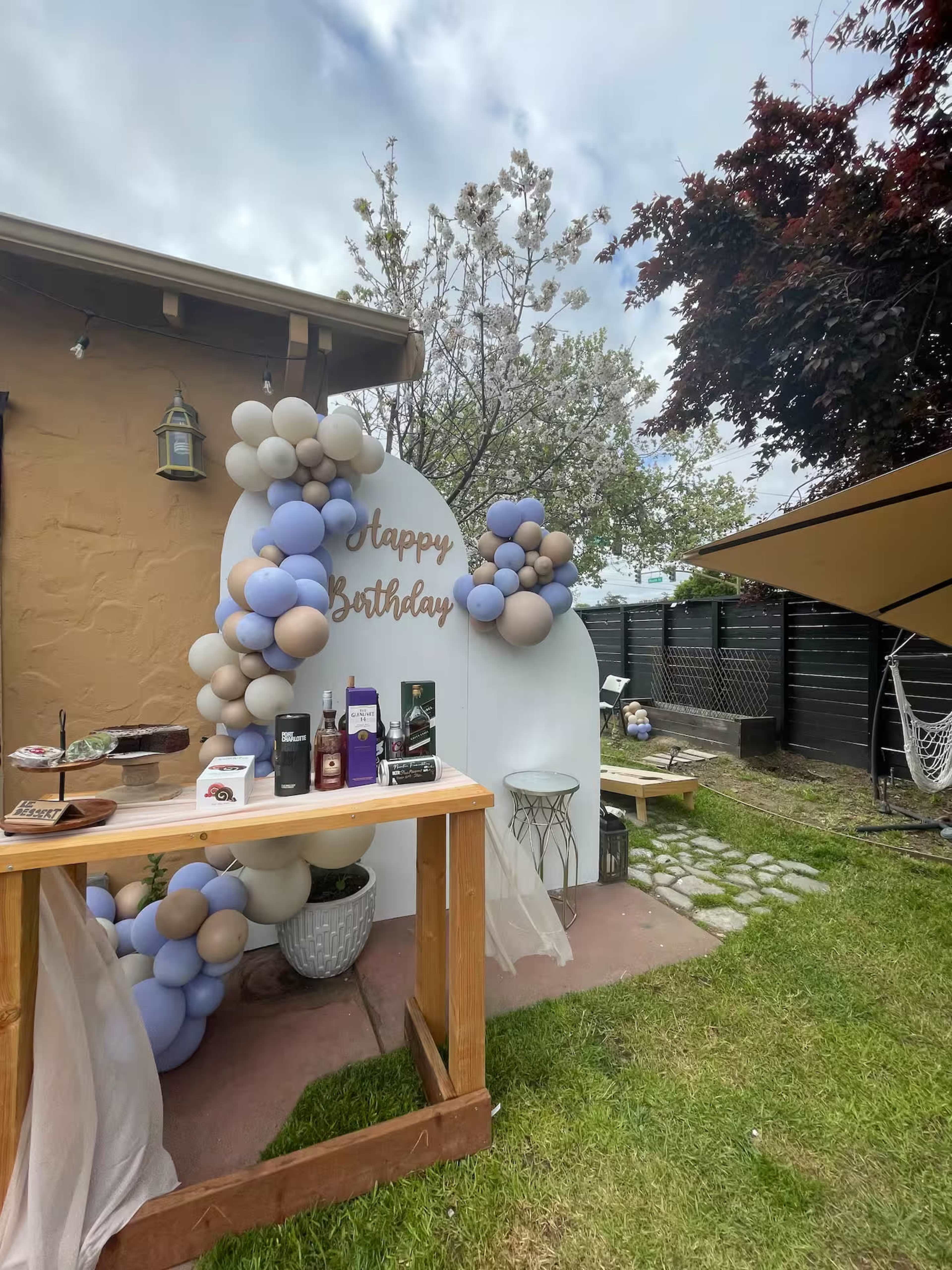 The image shows a birthday celebration setup in a backyard, featuring a decorated backdrop with balloons, a wooden table displaying drinks, and a lush green lawn.