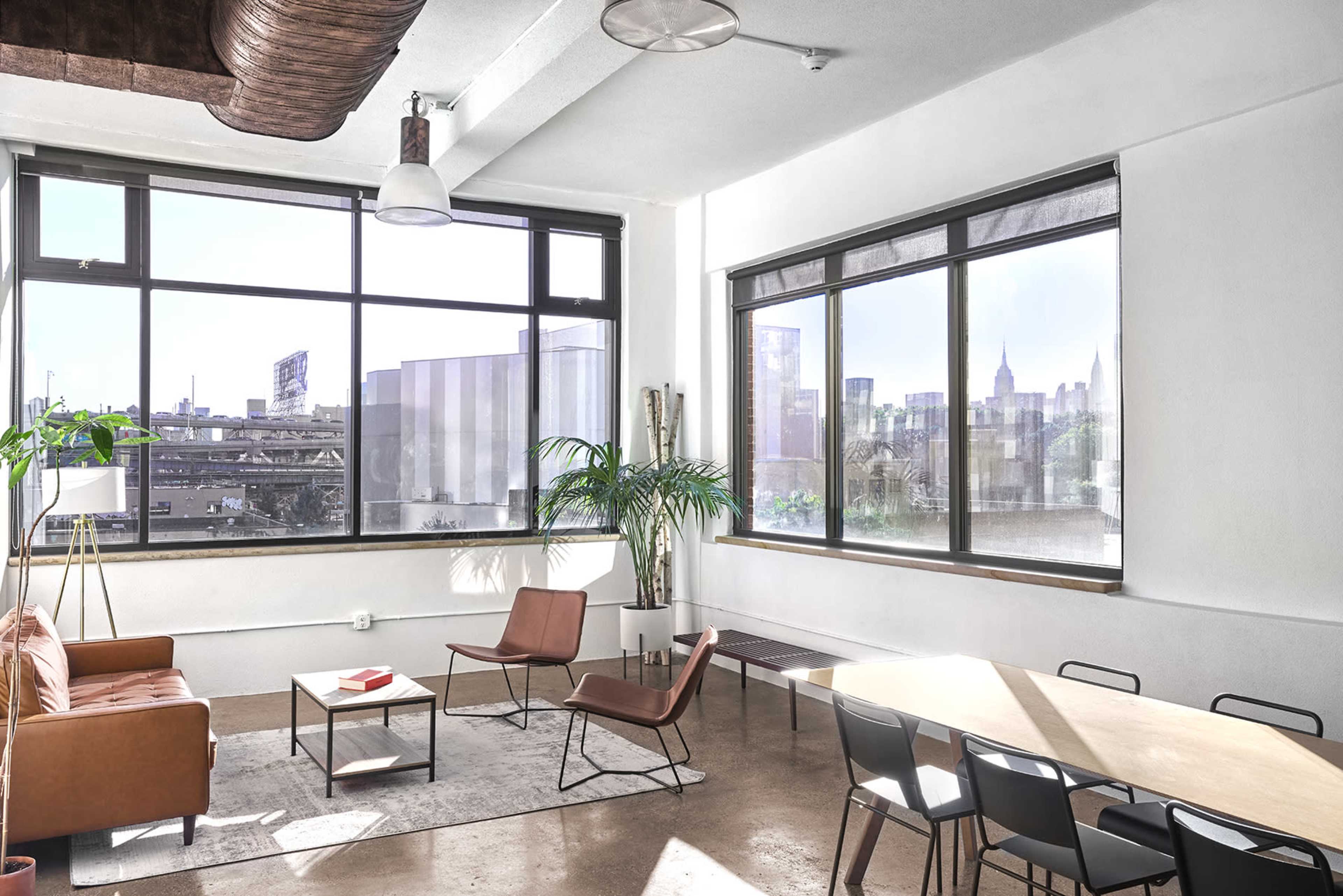 The image shows a modern room with large windows, a brown leather sofa, minimalist furniture, and a view of a cityscape through the glass.