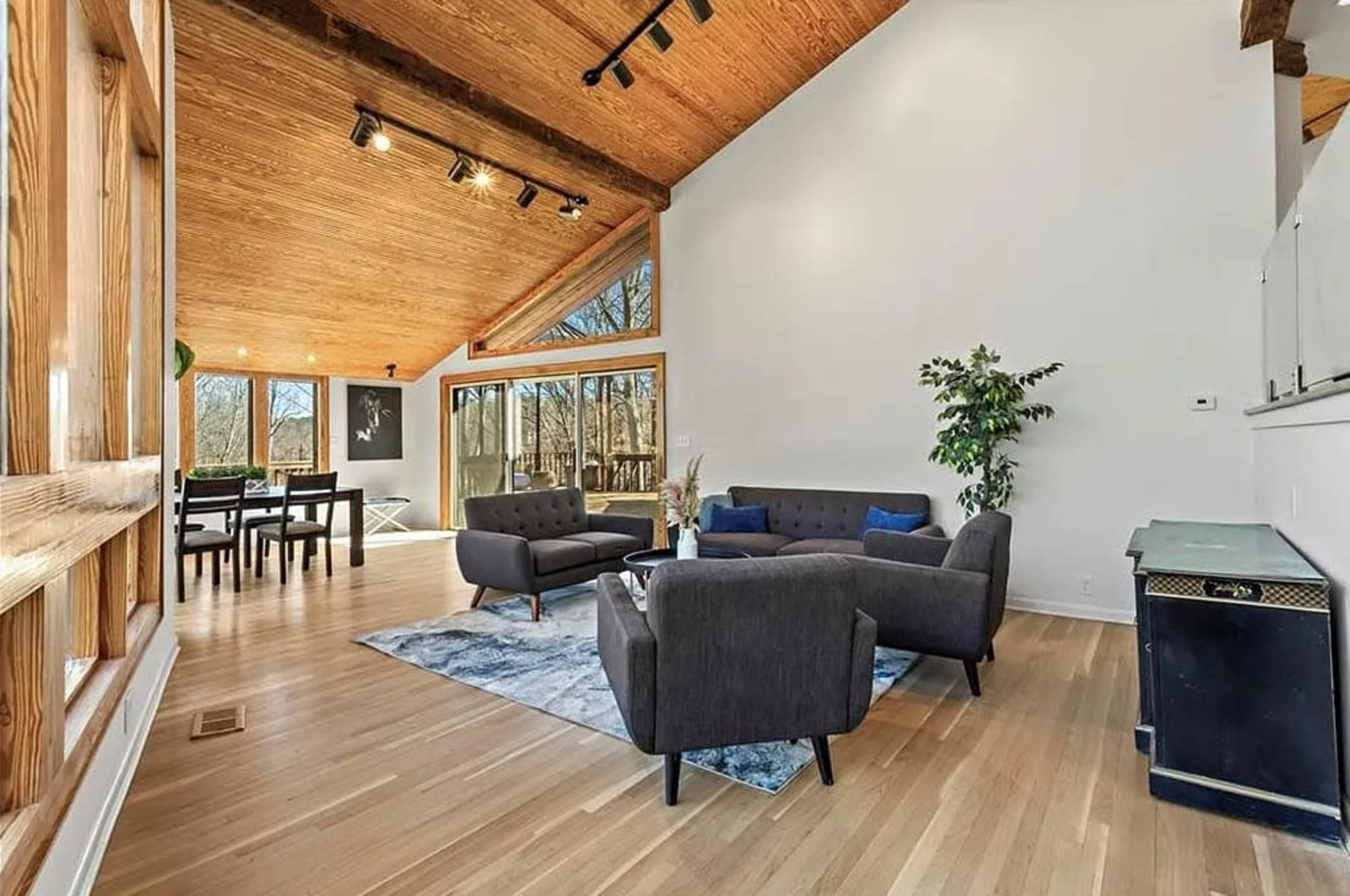 The image shows a spacious living room with large windows, wooden beams, a small dining area, and modern furniture arranged around a rug.