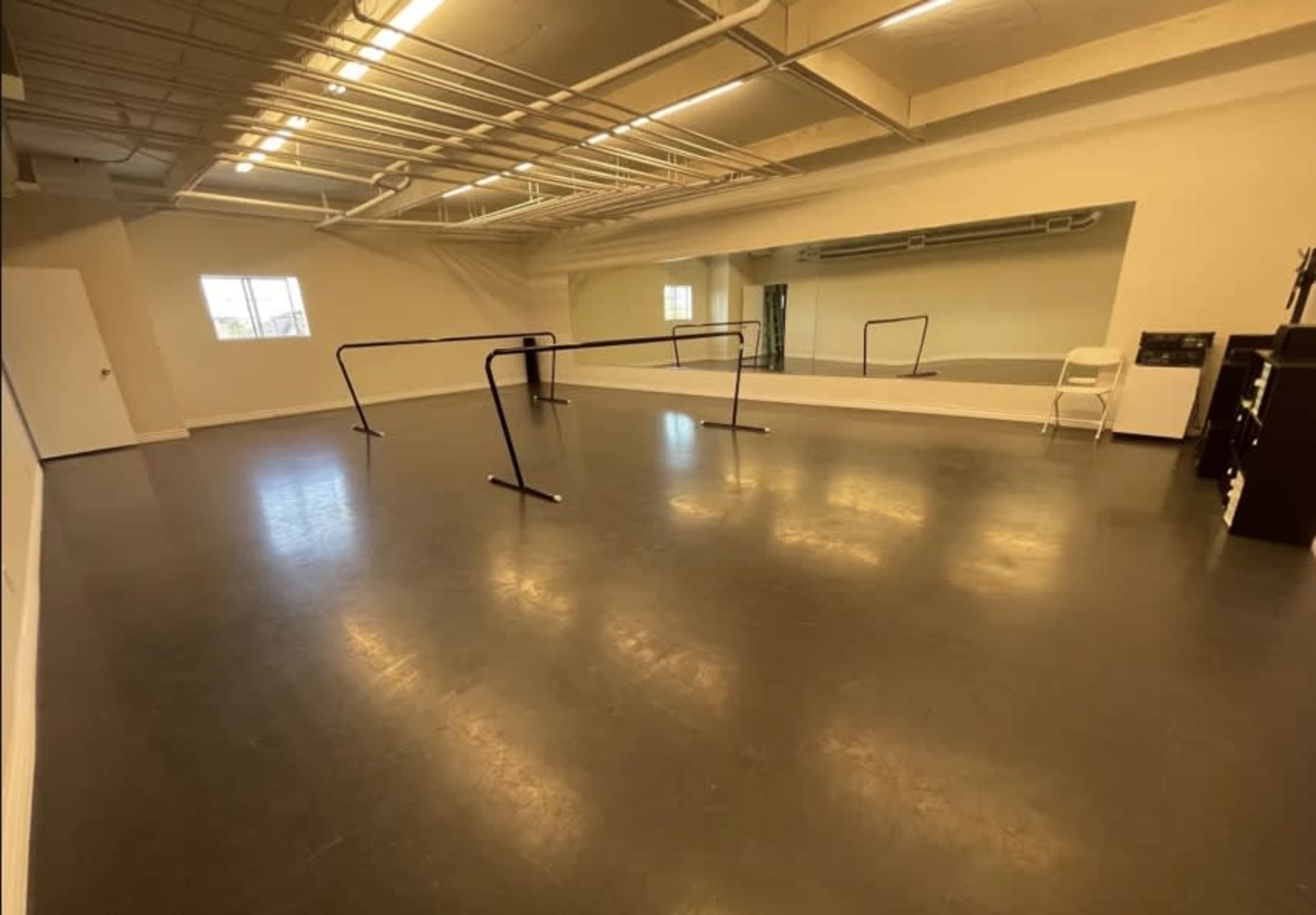 A spacious dance studio with a polished floor, barre equipment along one wall, and a large mirror reflecting the room.