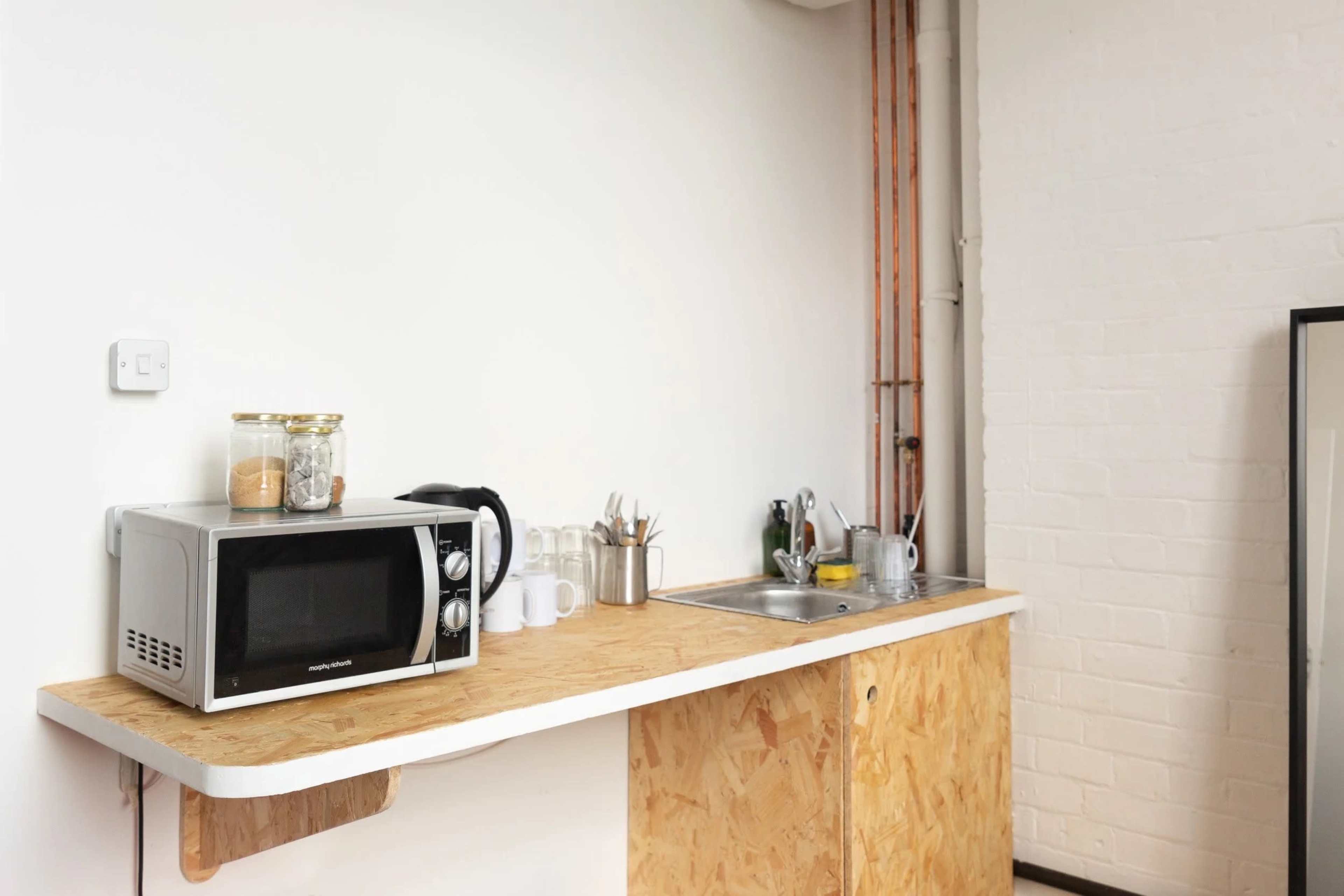 A small kitchen area features a microwave, a sink with dishes, and a countertop made of plywood.