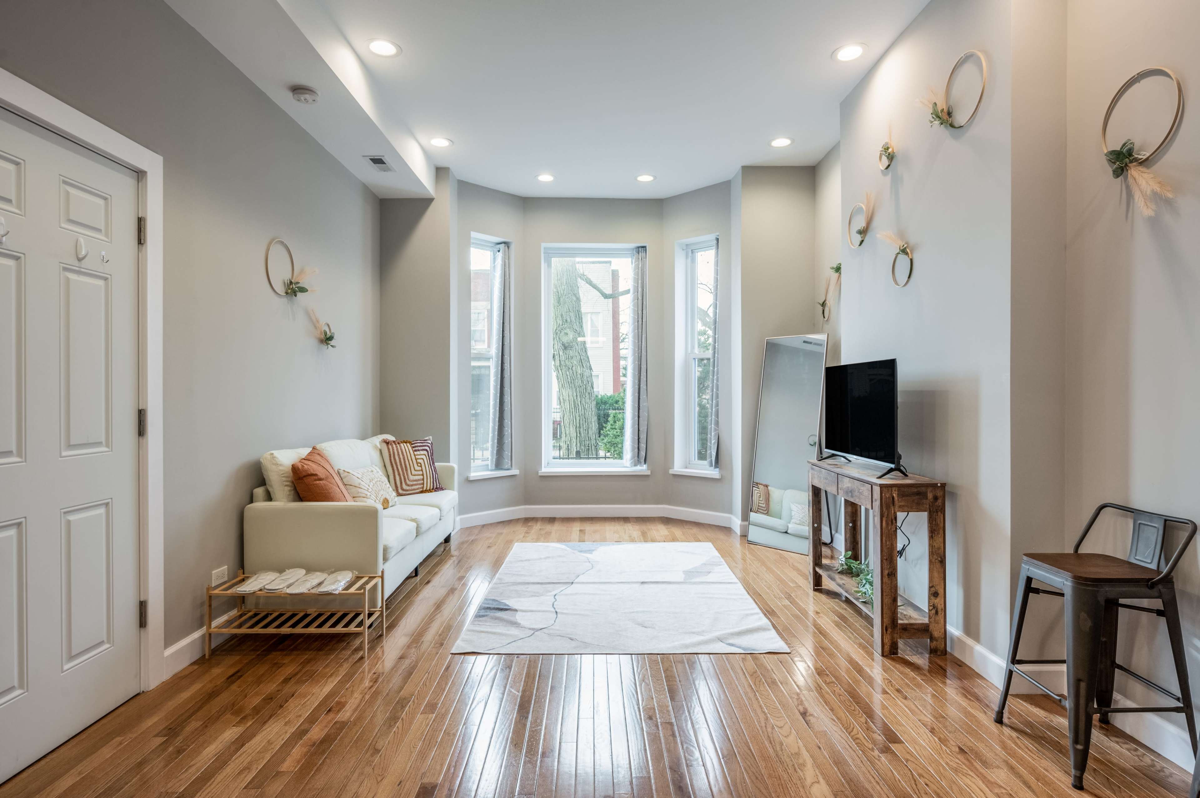 A modern living room with a light-colored sofa, a small wooden table, a wall-mounted TV, and large windows allowing natural light to enter.