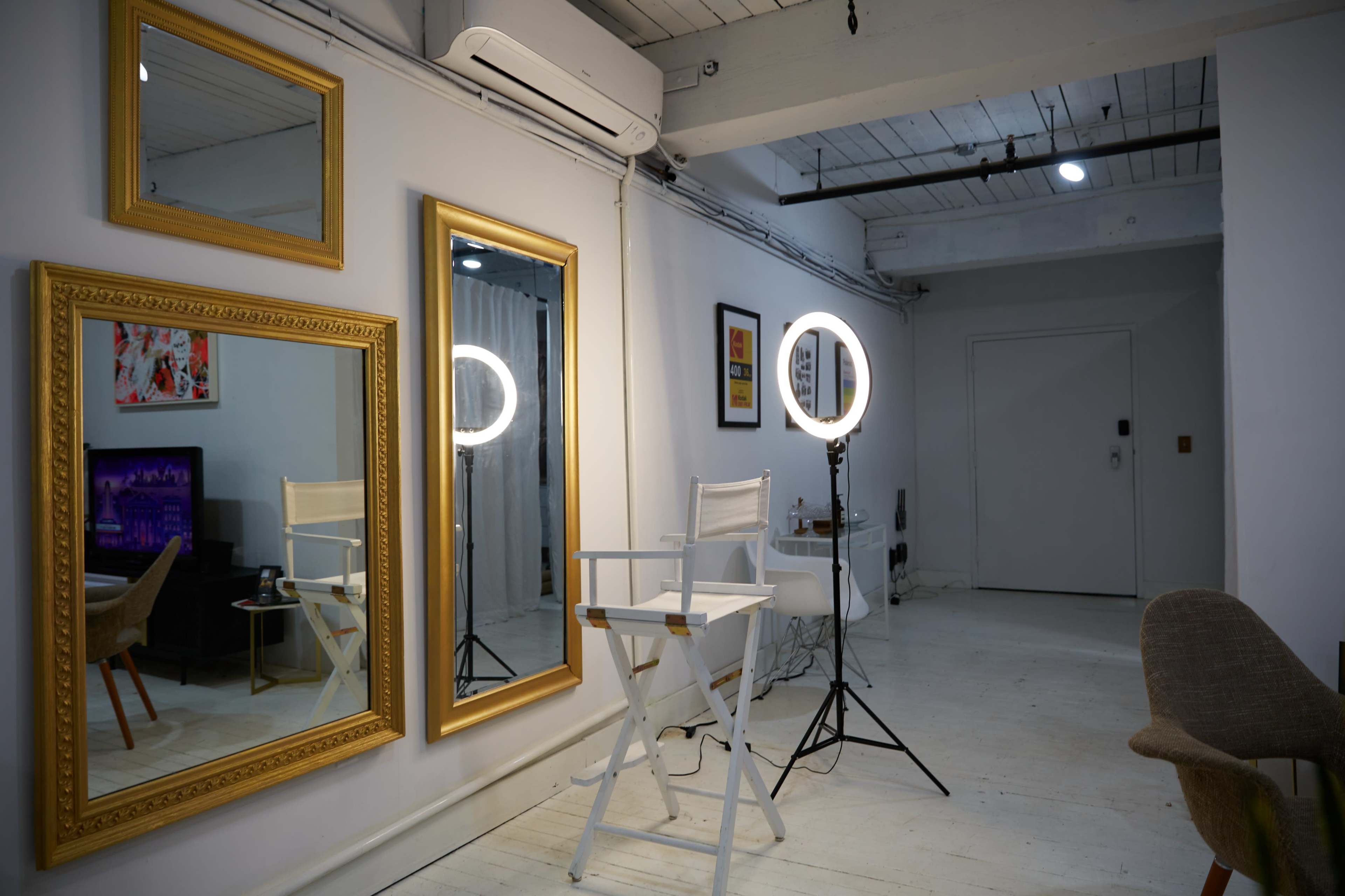 A well-lit room featuring two large mirrors, a ring light on a stand, and a tall director's chair against a white wall with wooden beams.