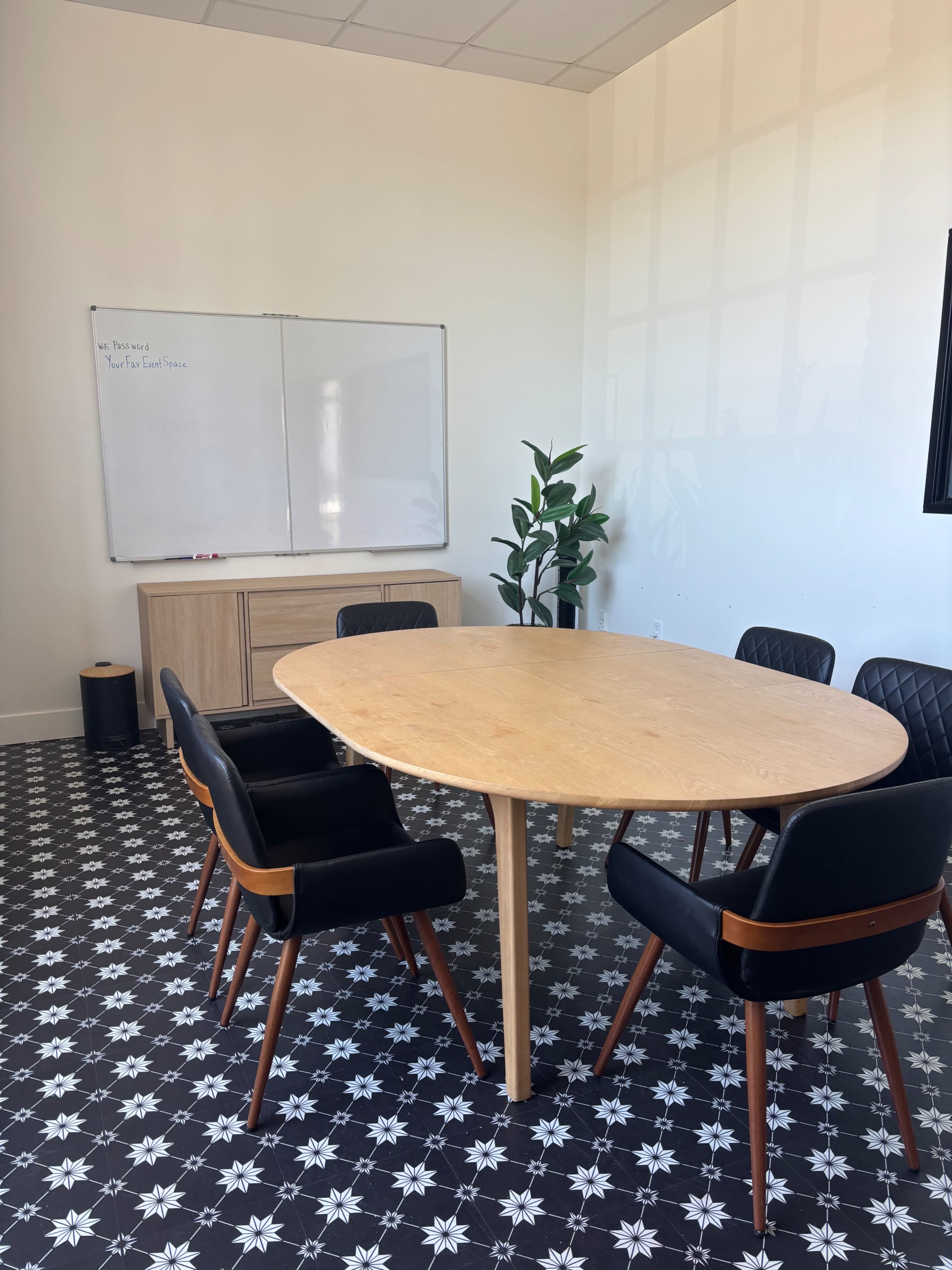 A meeting room features an oval wooden table surrounded by black chairs, with a whiteboard and a tall plant in the background.