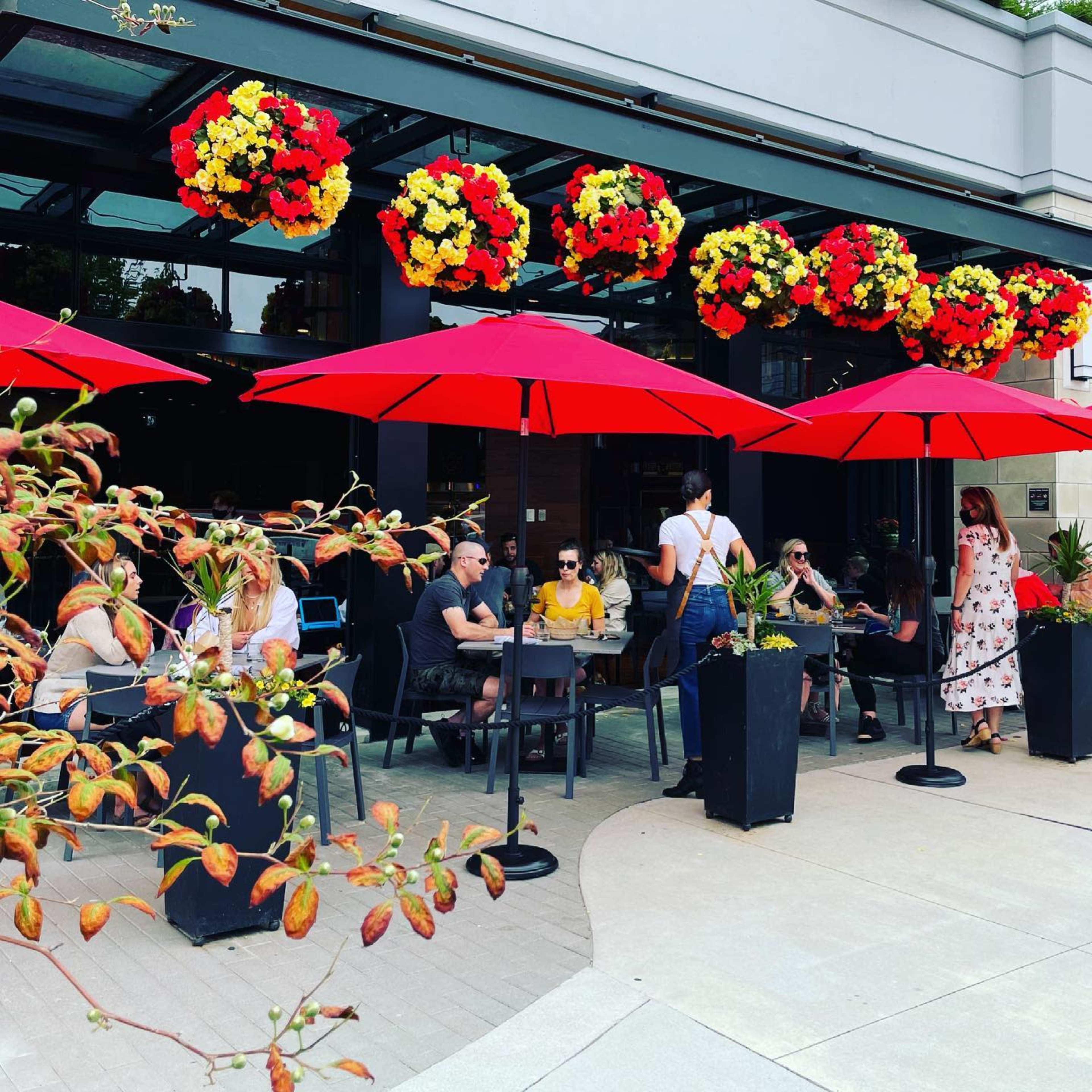 A group of people is seated at tables outside a restaurant, with red umbrellas and vibrant flower arrangements overhead.