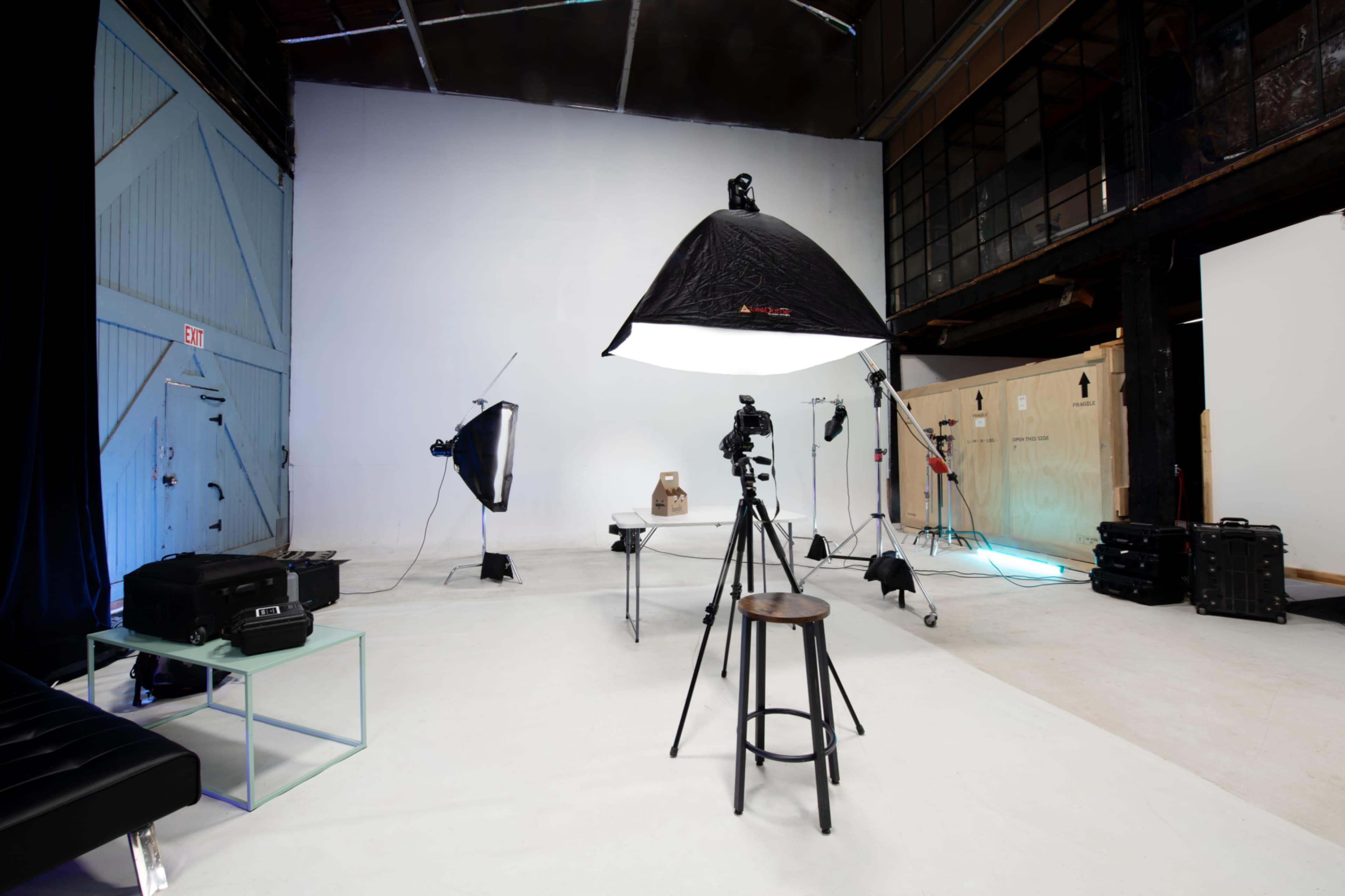 The image shows a photography studio setup with various lighting equipment, a camera on a tripod, and a minimalist white backdrop.
