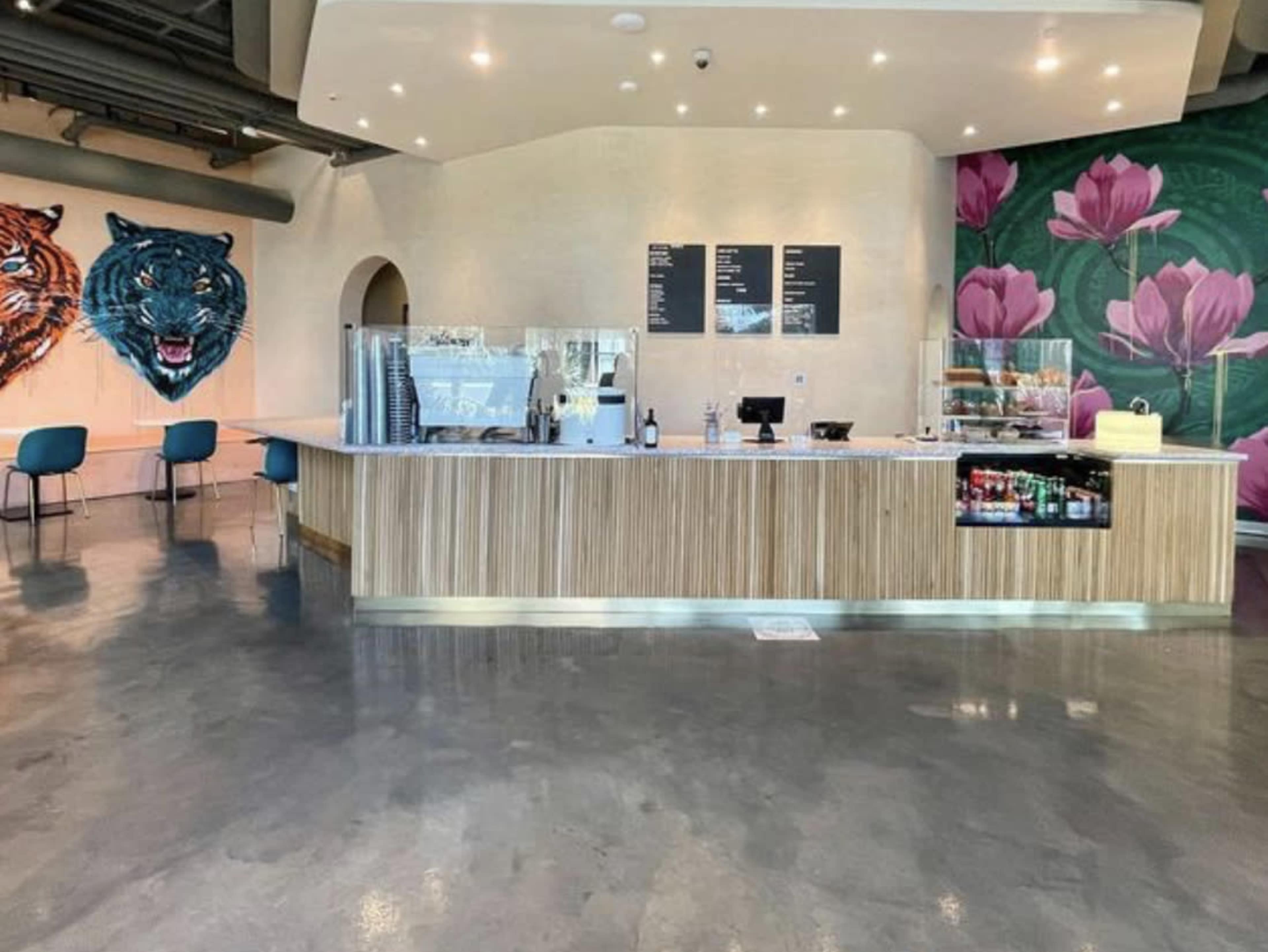A modern cafe with a wooden counter, a display of pastries, and colorful murals of a tiger and lotus flowers on the walls.