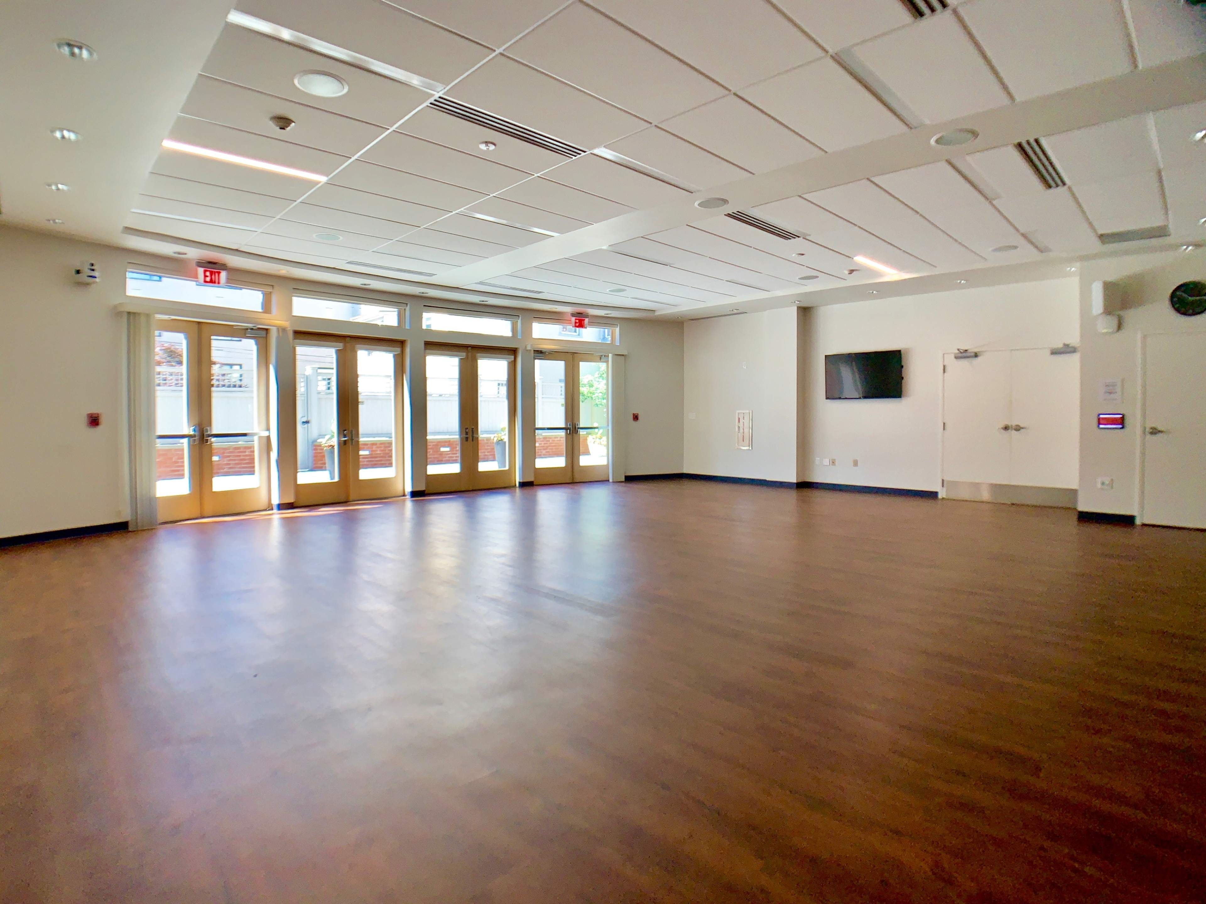 A spacious, empty room with wood flooring, large windows, and double doors leading outside.