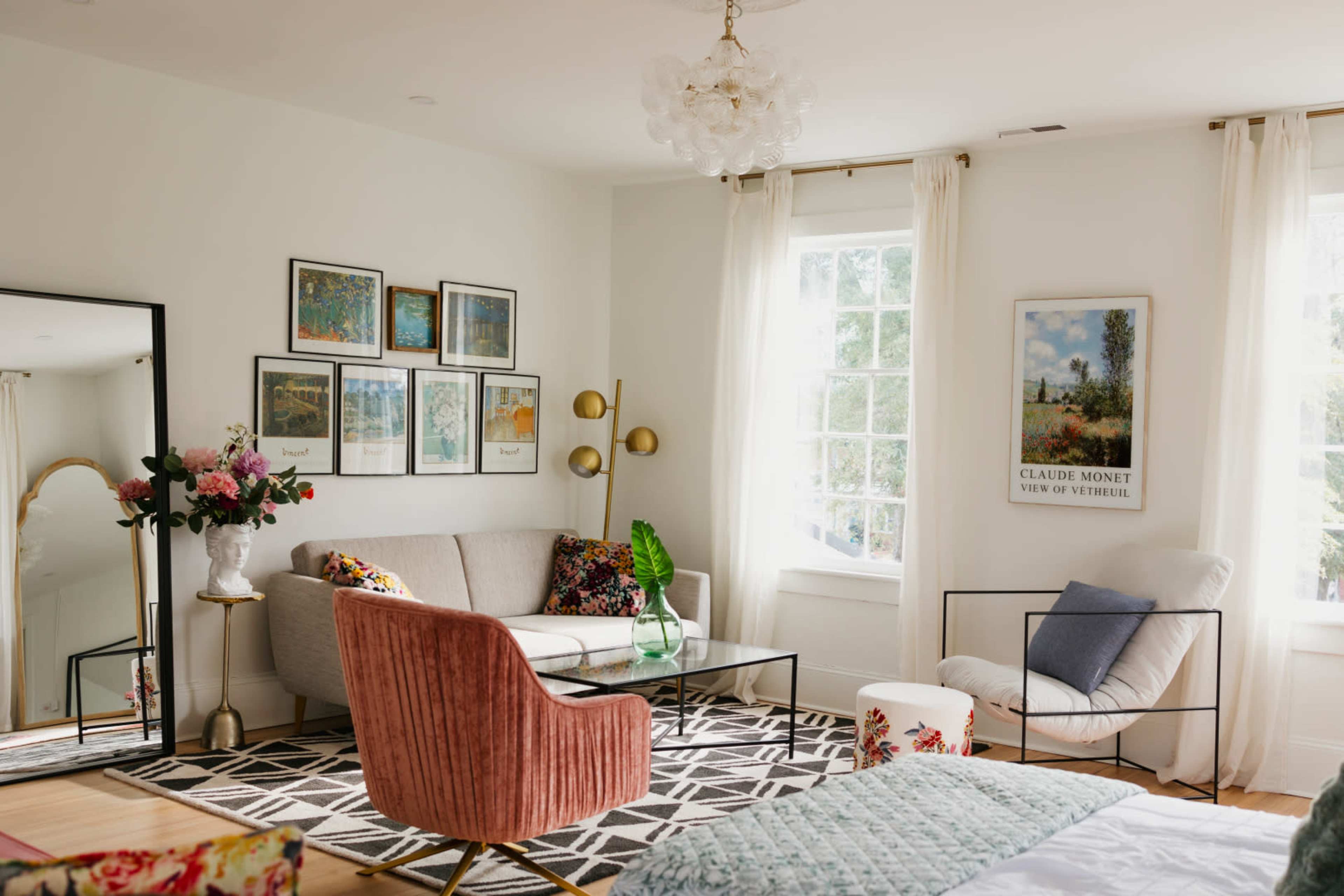 The image shows a stylish living room with a variety of framed artworks on the wall, a gray sofa, a pink armchair, and a coffee table, all arranged around a patterned rug.