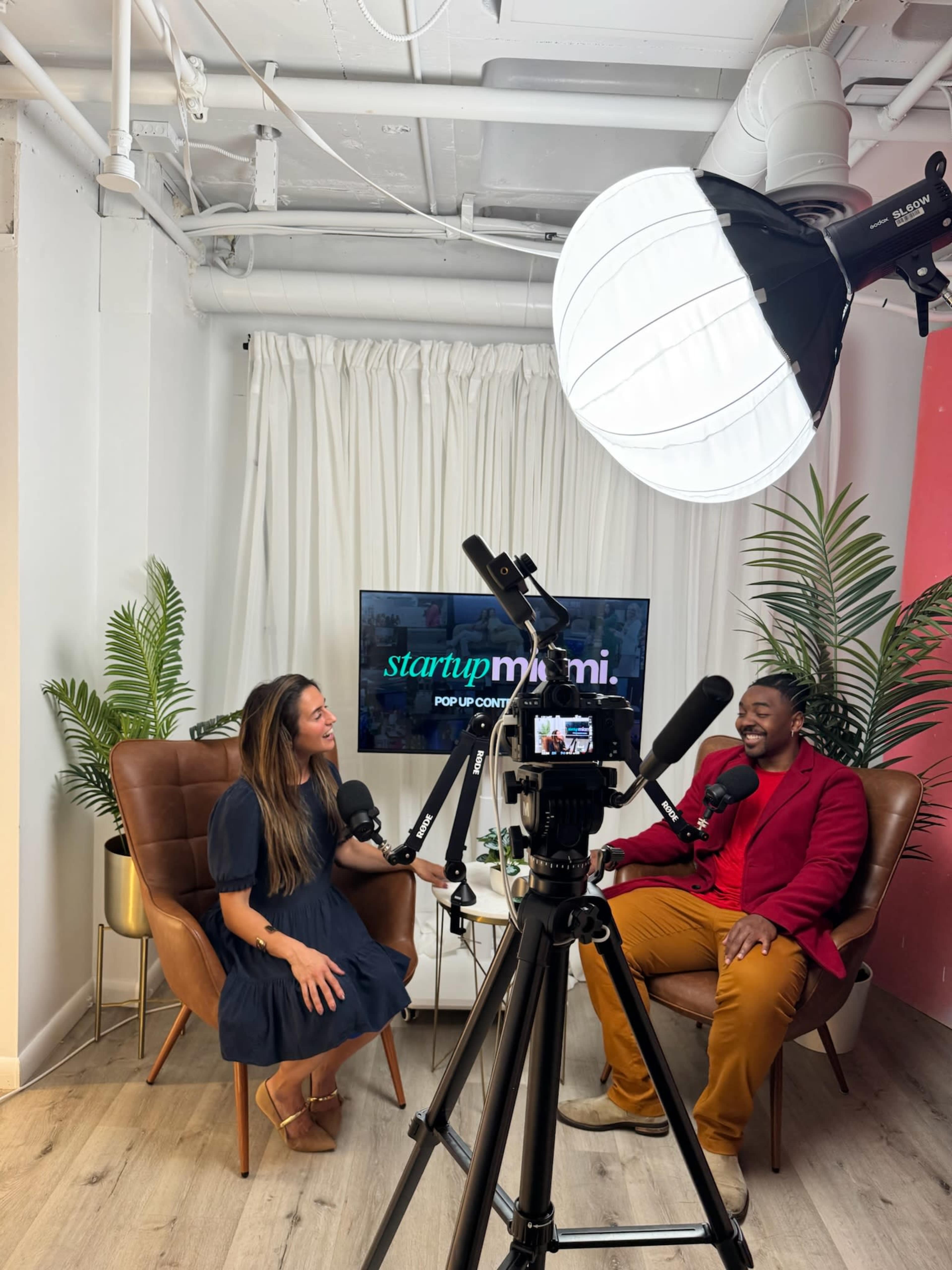 A woman and a man sit in a studio setting with a camera and microphone in front of them, and a screen behind them displays the text "startup miami."