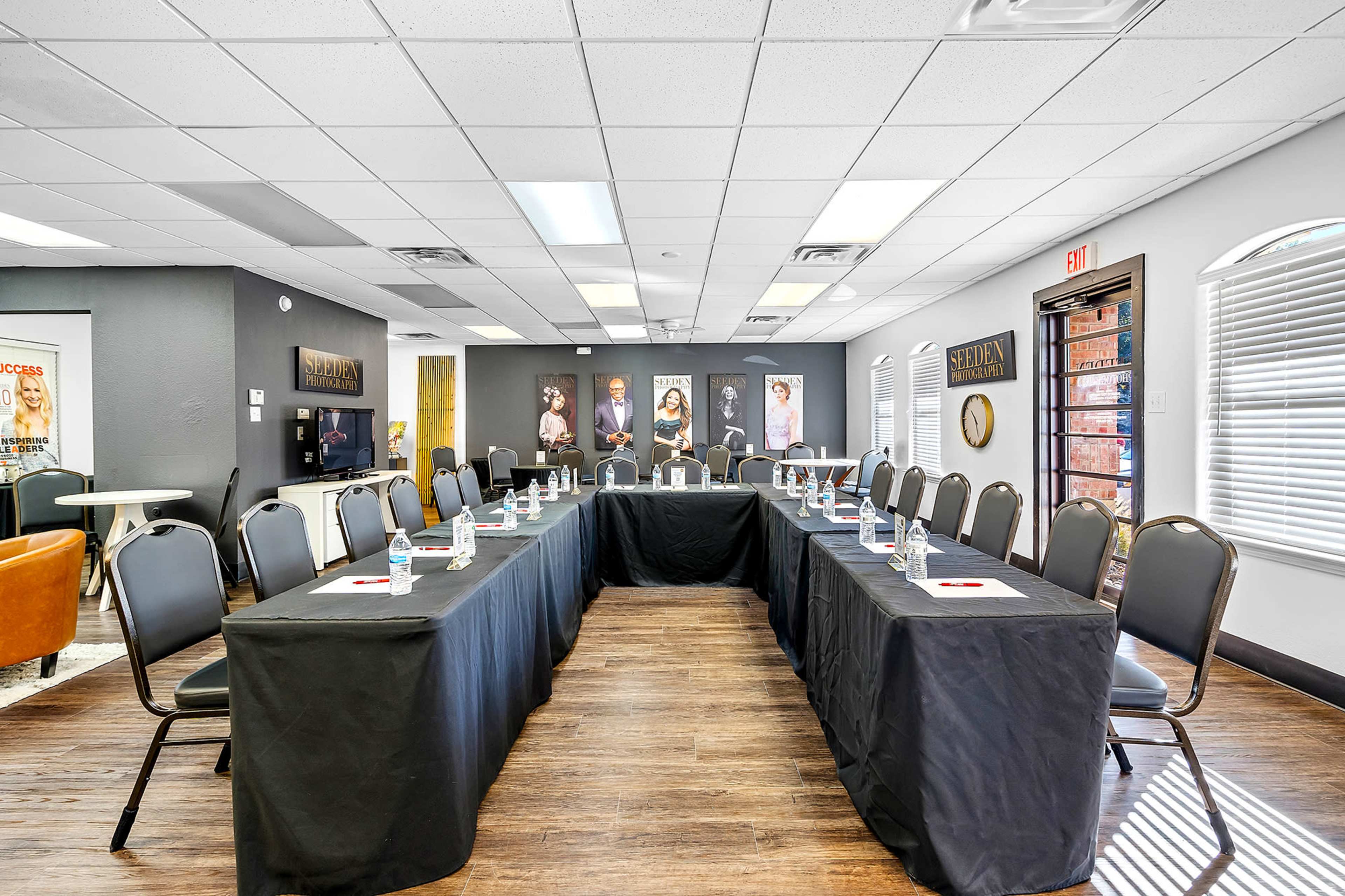 A conference room is set up with two rectangular tables arranged in a U-shape, each covered with black tablecloths and surrounded by chairs, with water bottles placed at each seat.