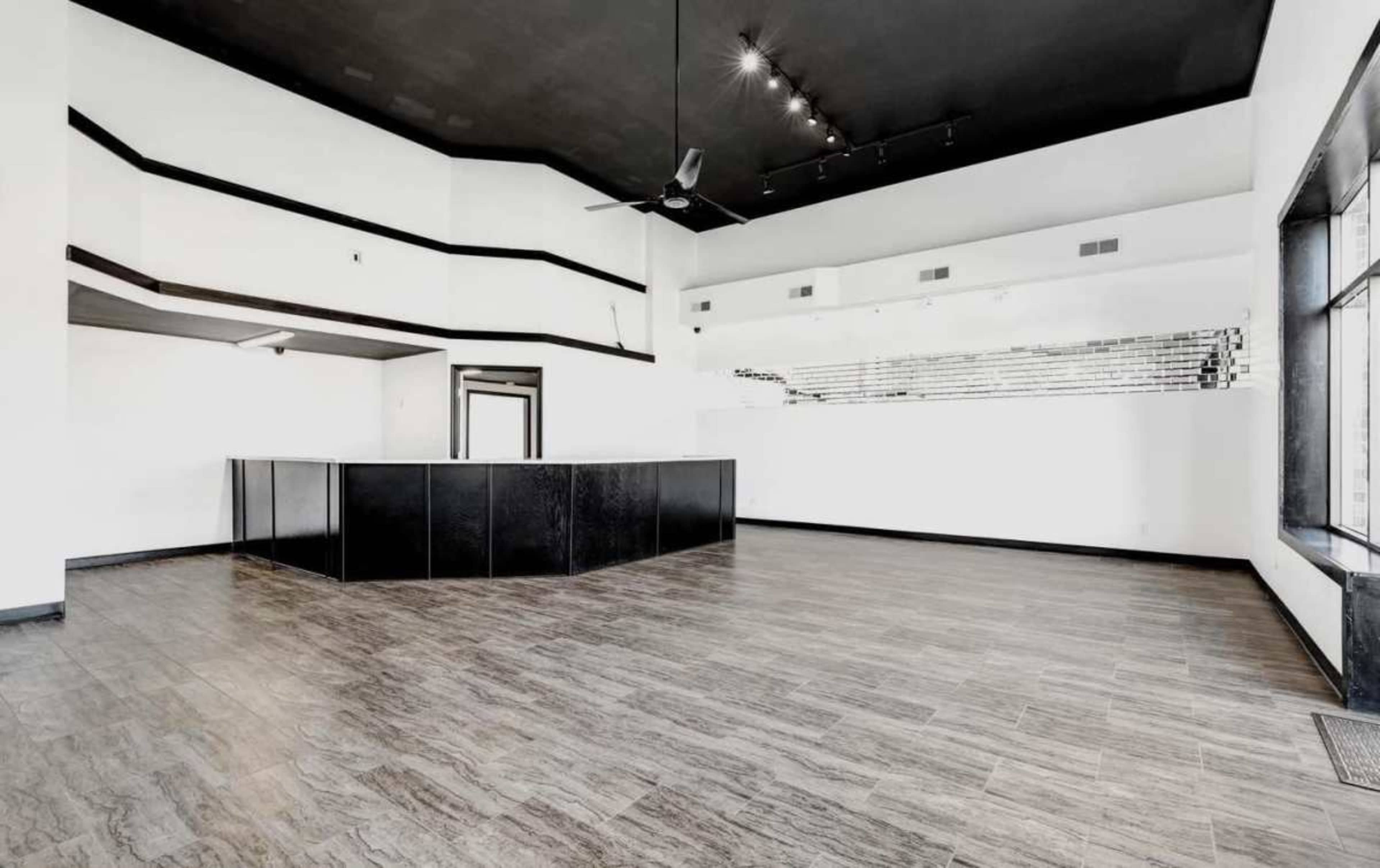 The image shows an empty commercial space featuring a central black reception counter, wooden flooring, and white walls with large windows.