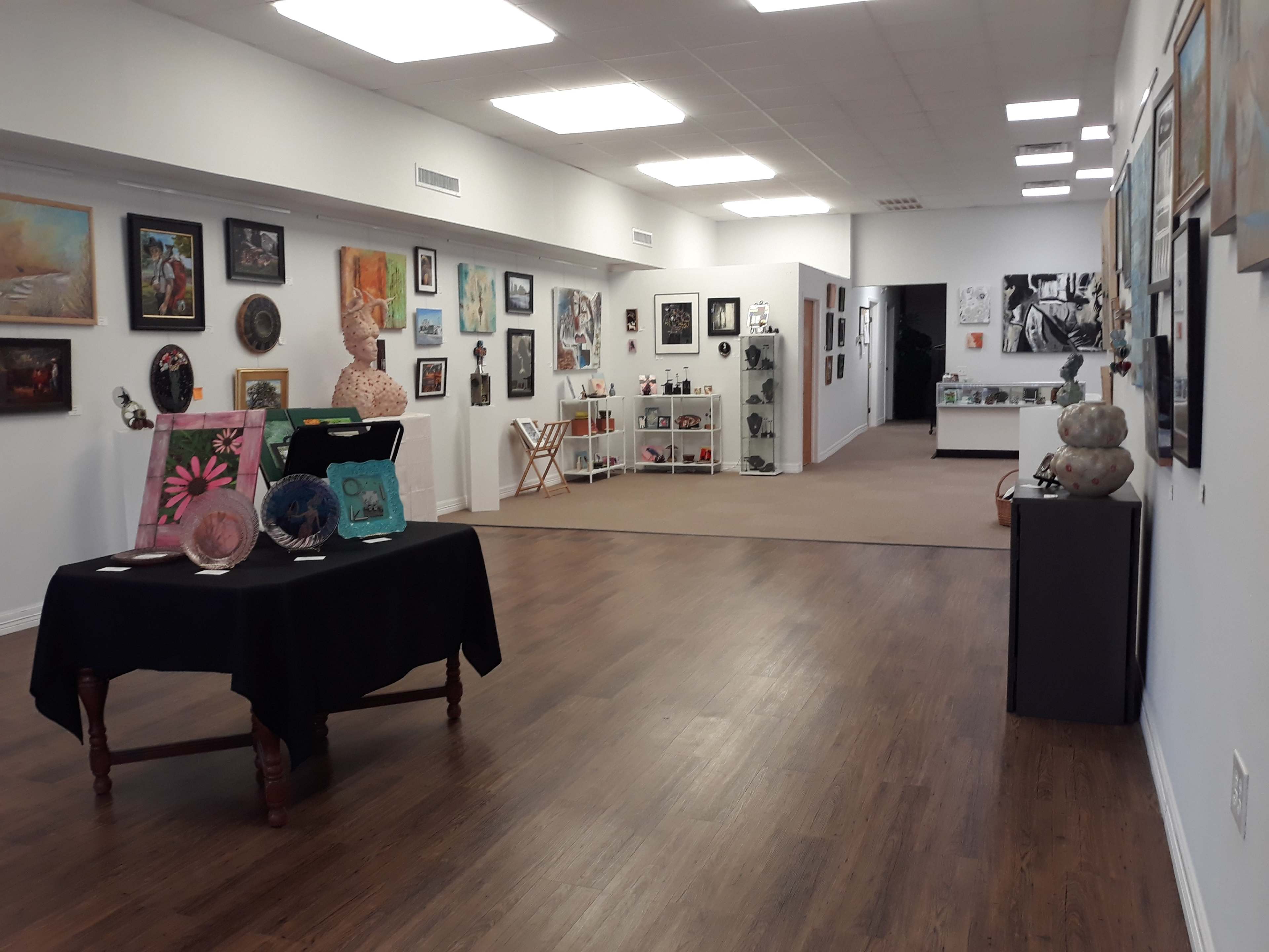 An art gallery features various paintings and sculptures displayed on the walls and tables, with a wooden floor and bright lighting.