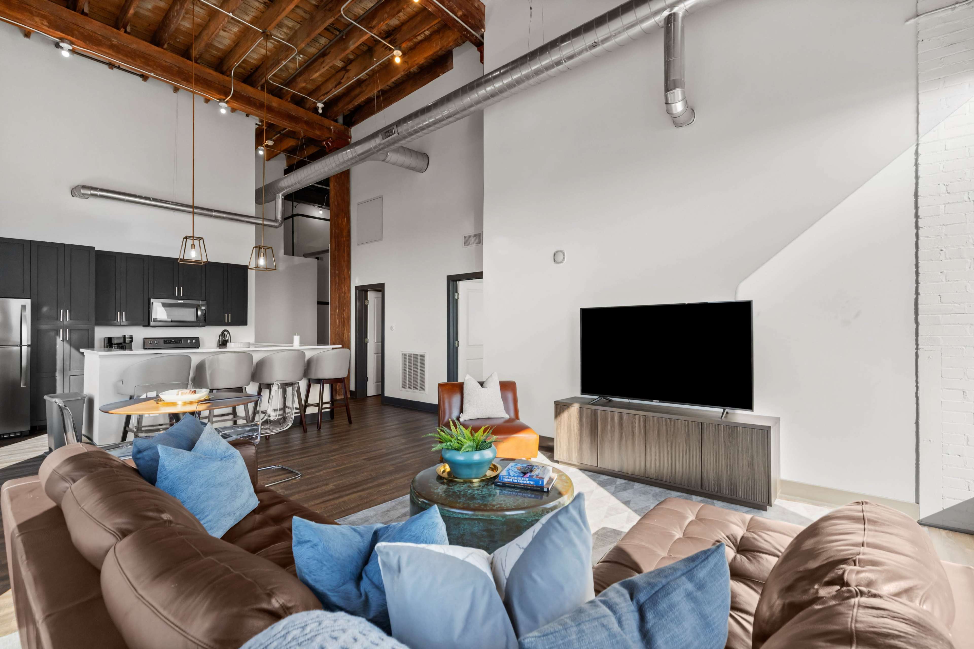 The image shows a modern, open-concept living area with a sectional sofa, a coffee table, and a television, alongside a kitchen featuring a breakfast bar and stainless steel appliances.