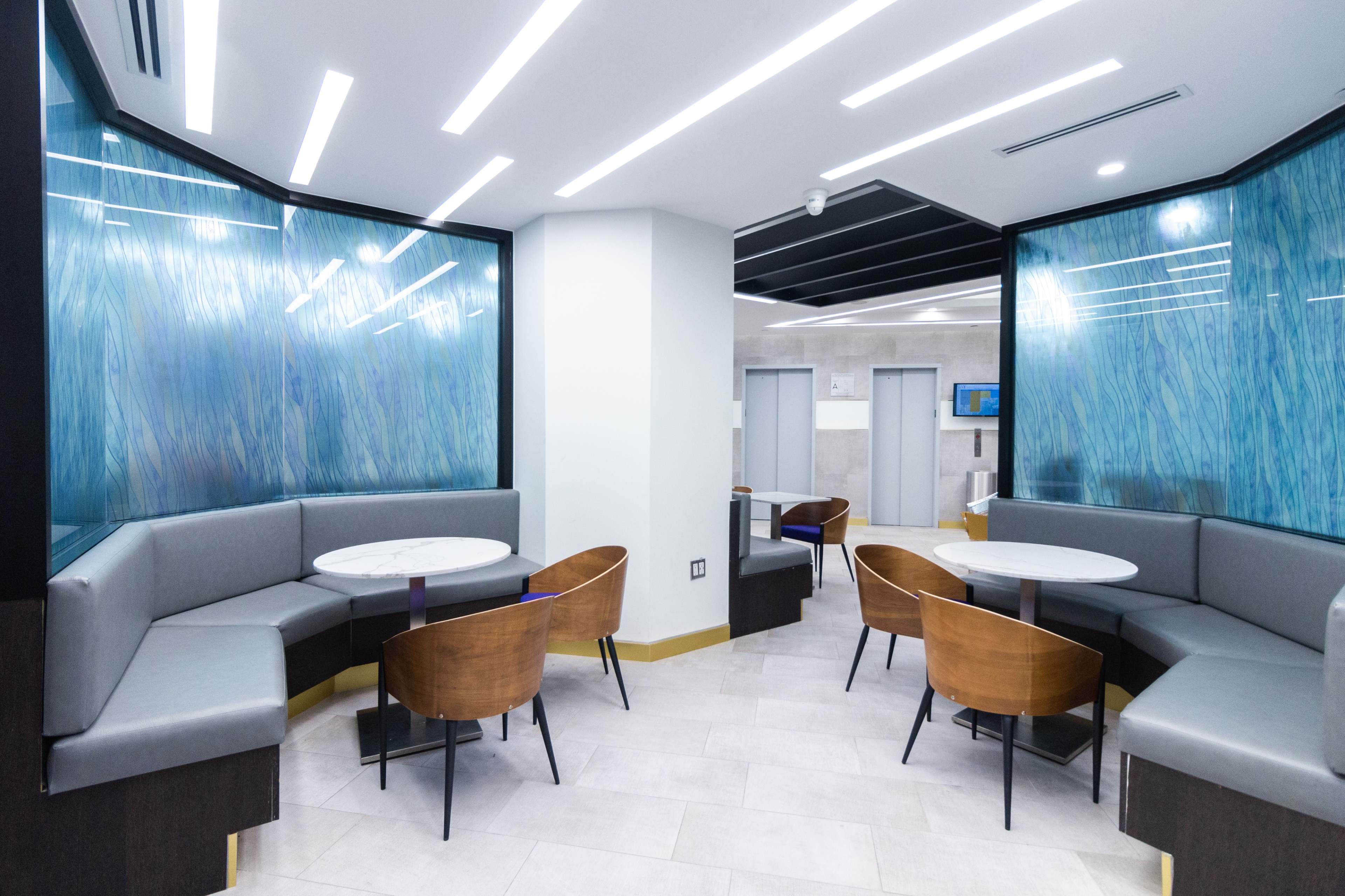 The image shows a modern waiting area with gray upholstered seating, circular marble tables, and blue glass panels.