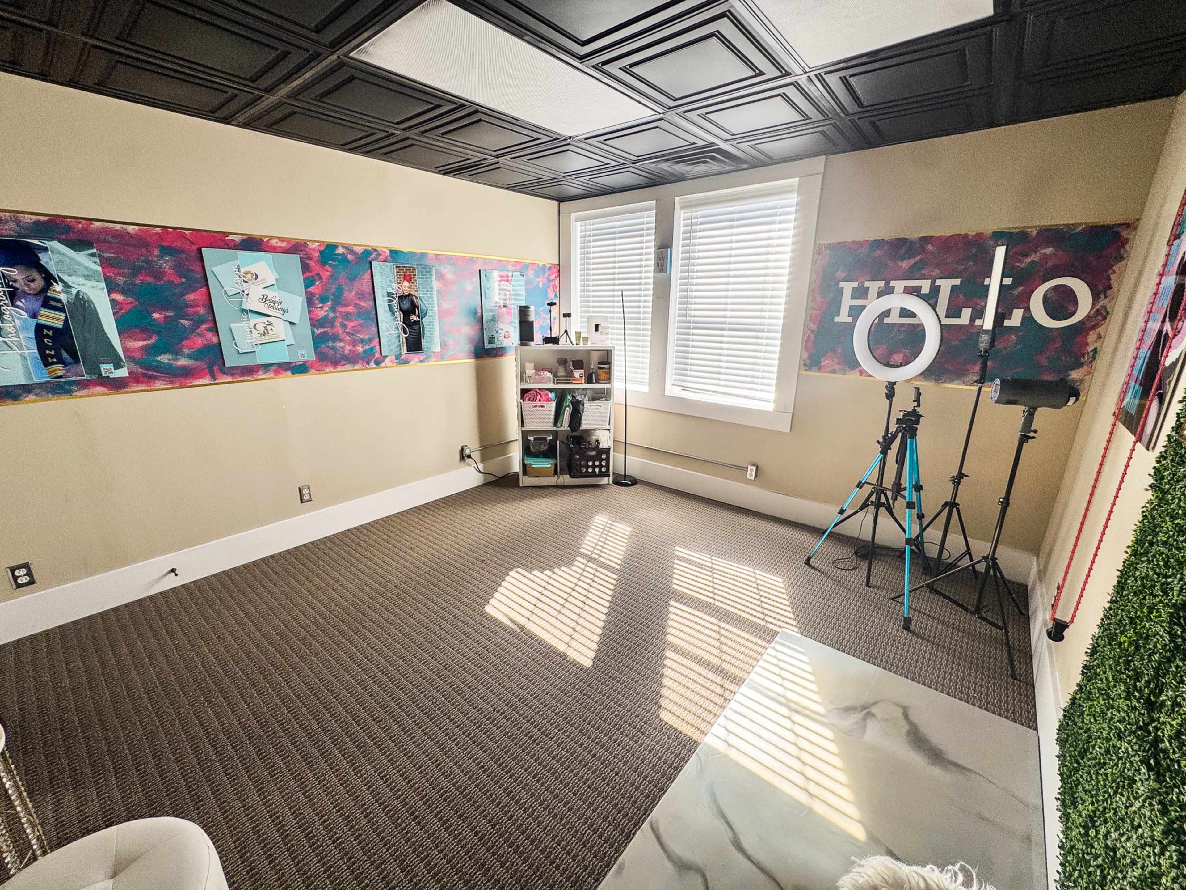 The image depicts a brightly lit room with patterned wall decorations, a shelving unit in the center, and a camera setup for photography or video on a tripod.