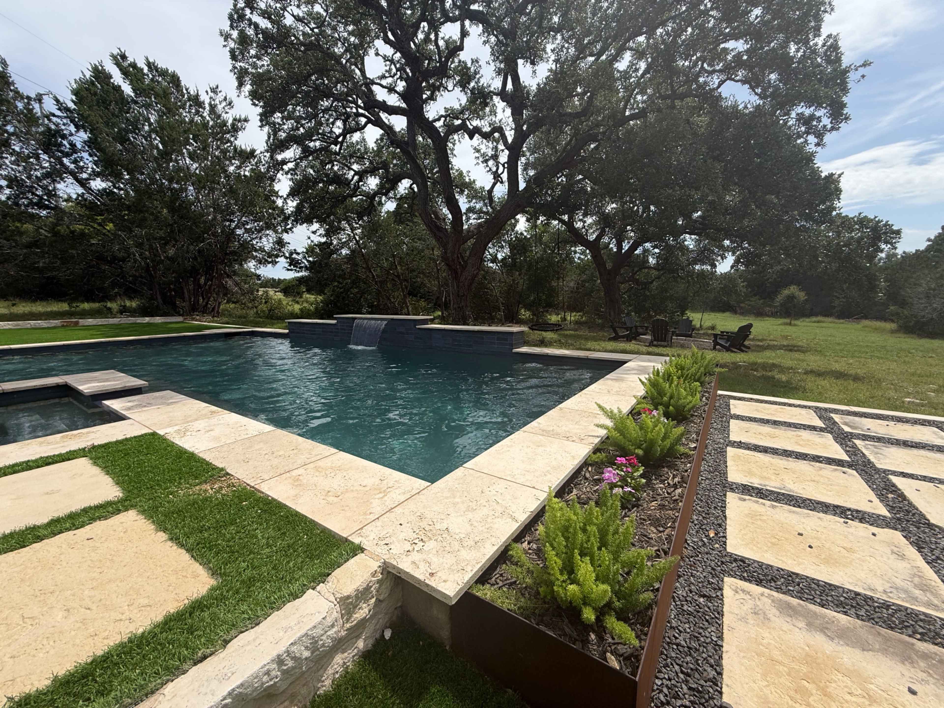 A rectangular pool with a water feature is surrounded by landscaped stone paving and greenery under a clear sky.