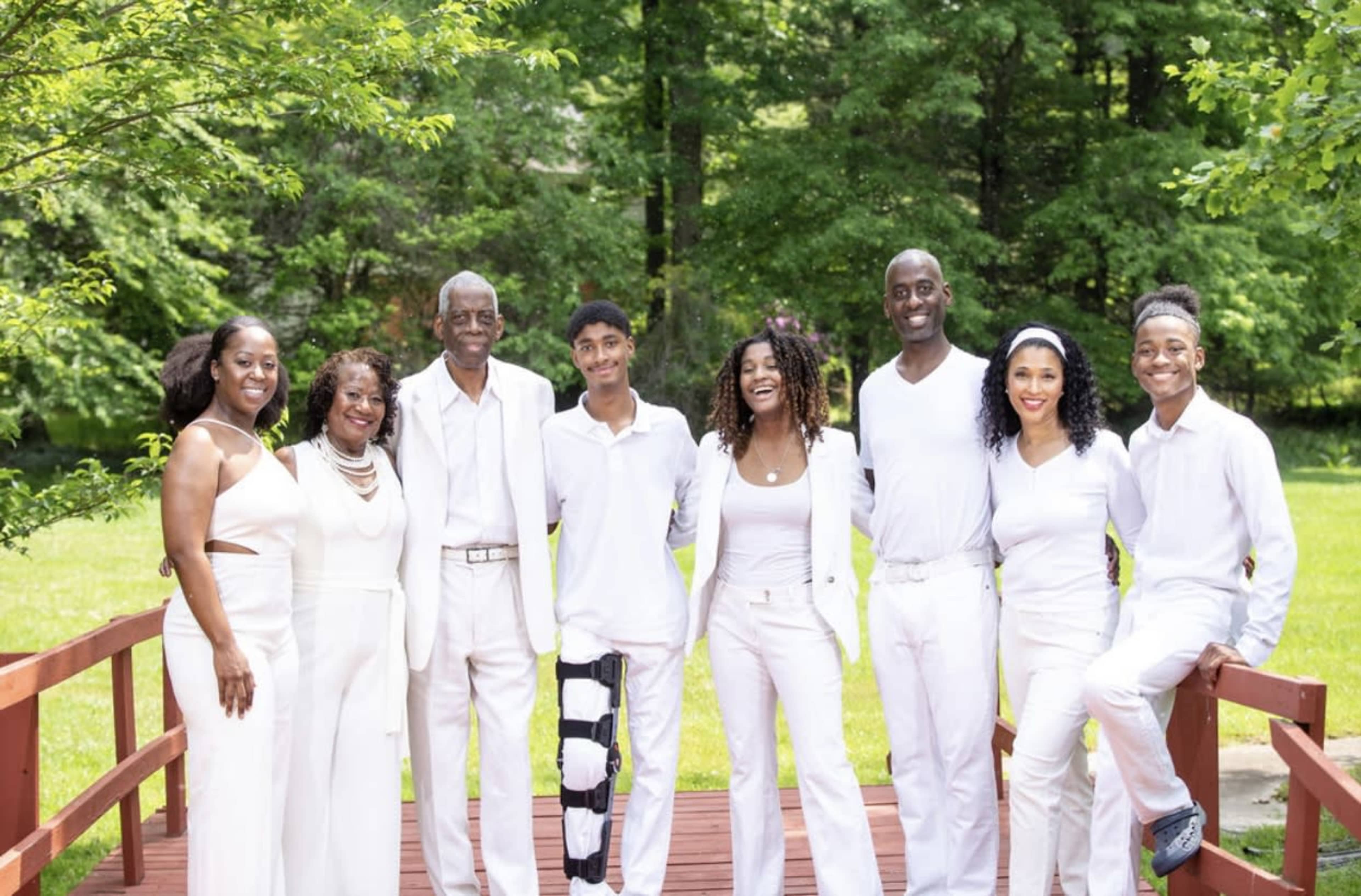 A family group dressed in white poses together on a wooden bridge in a green, outdoor setting.