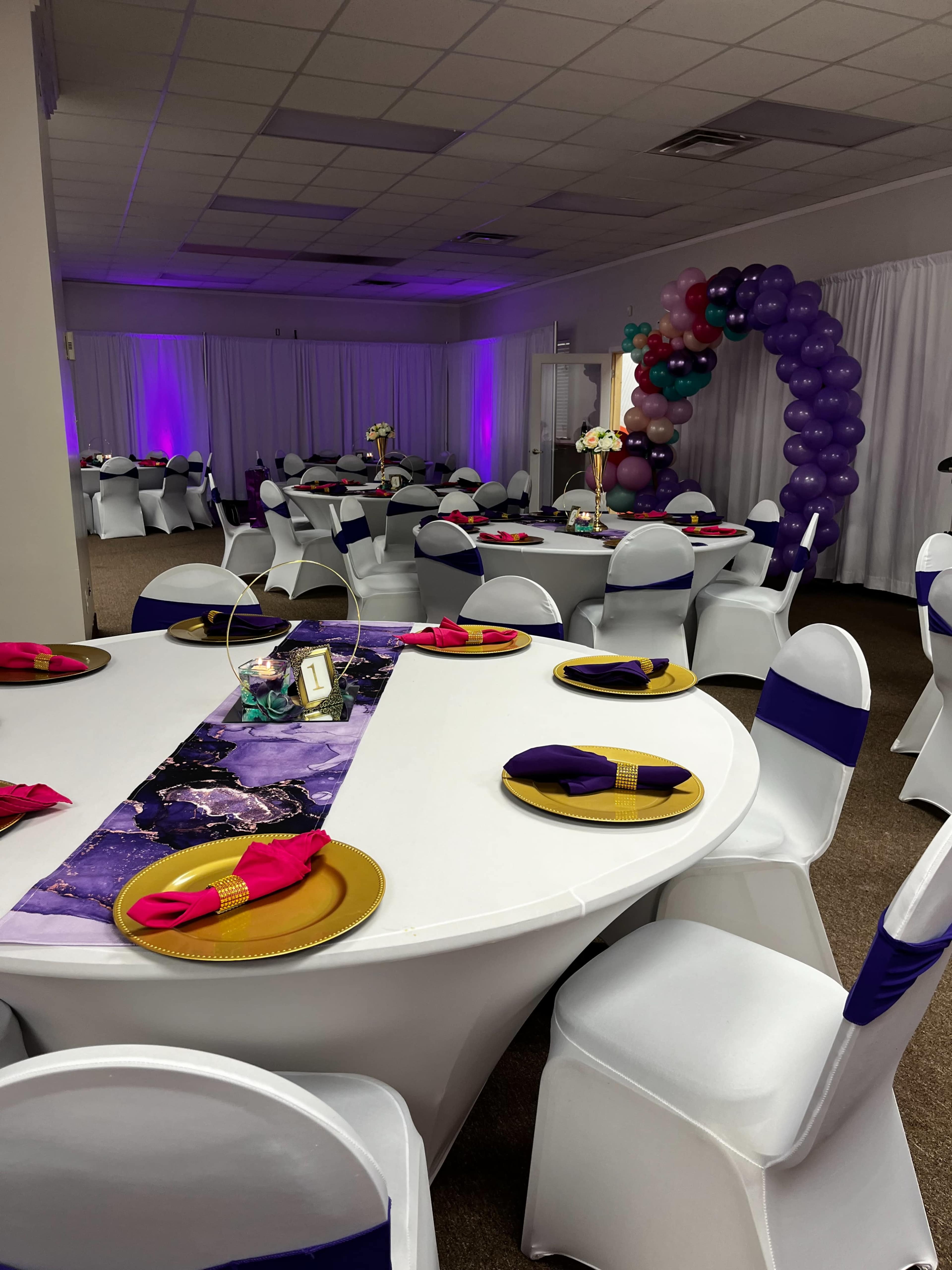 A decorated event space with tables arranged for a gathering, featuring white tablecloths, colorful napkins, and a balloon arch in the background.