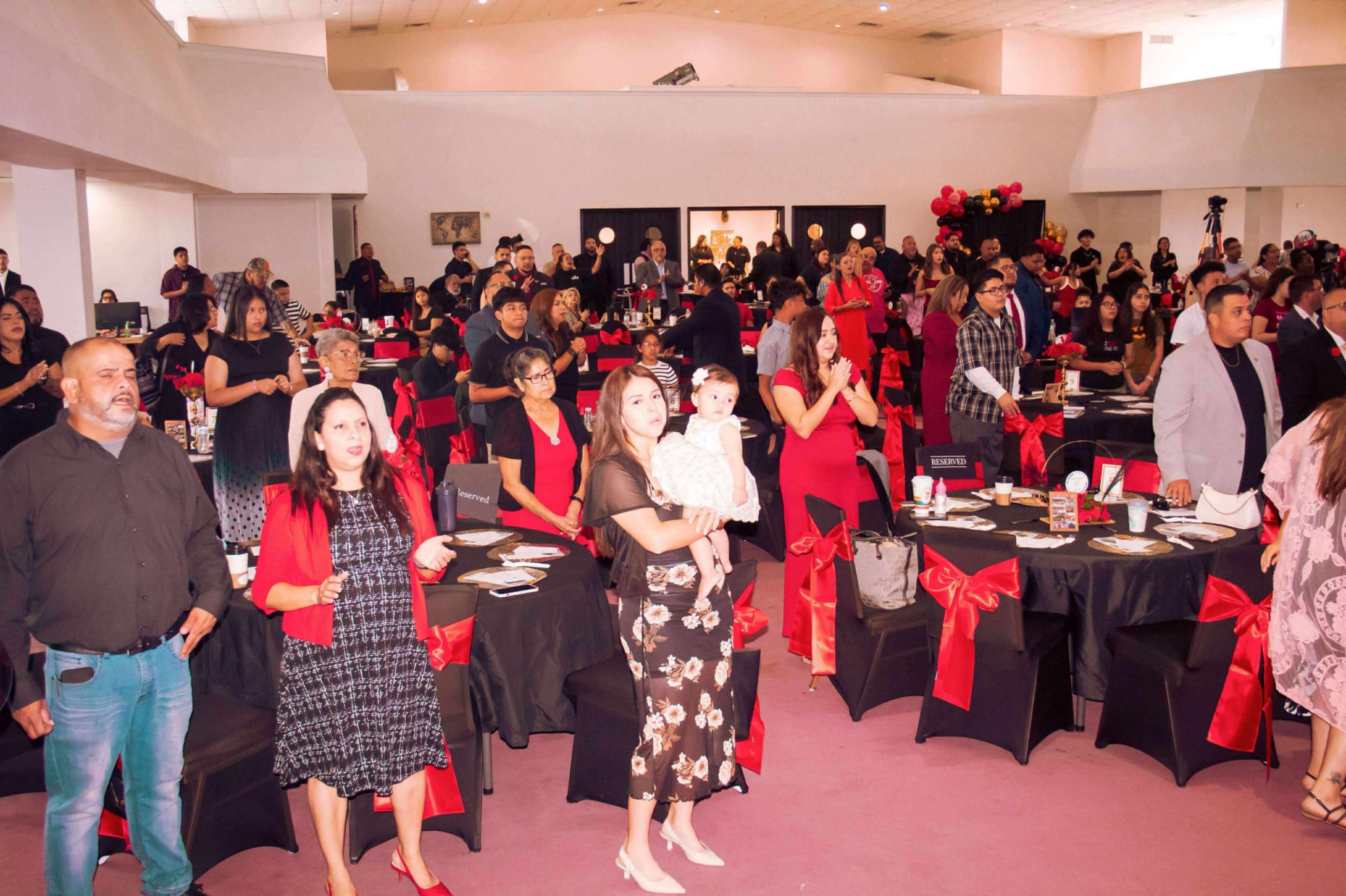 A large gathering of people dressed in formal attire is taking place in a spacious room, with tables adorned with black linens and red ribbon decorations.