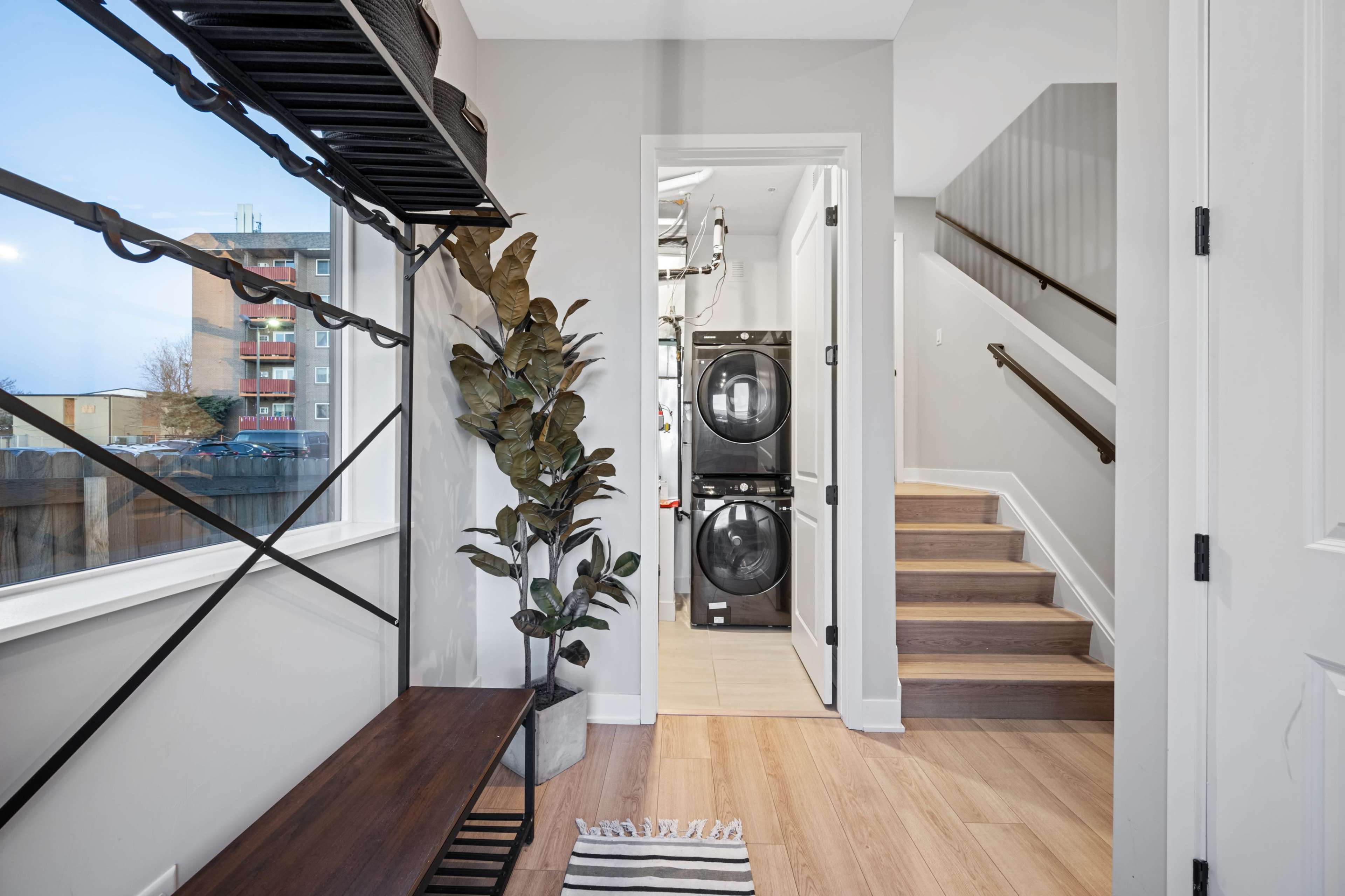 The image shows a hallway with a bench, a potted plant, and a staircase, leading to a laundry area with stacked washing machines.