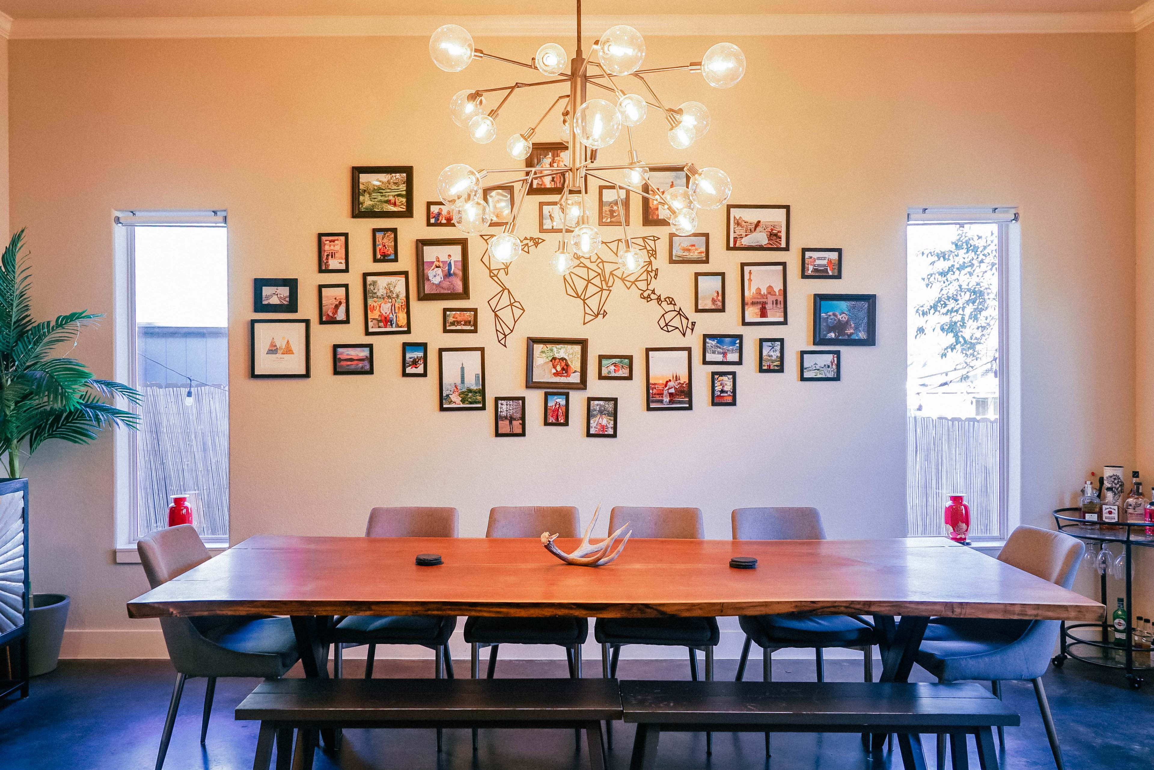A long wooden dining table with eight chairs is set against a wall adorned with multiple framed photographs and illuminated by a large modern chandelier.