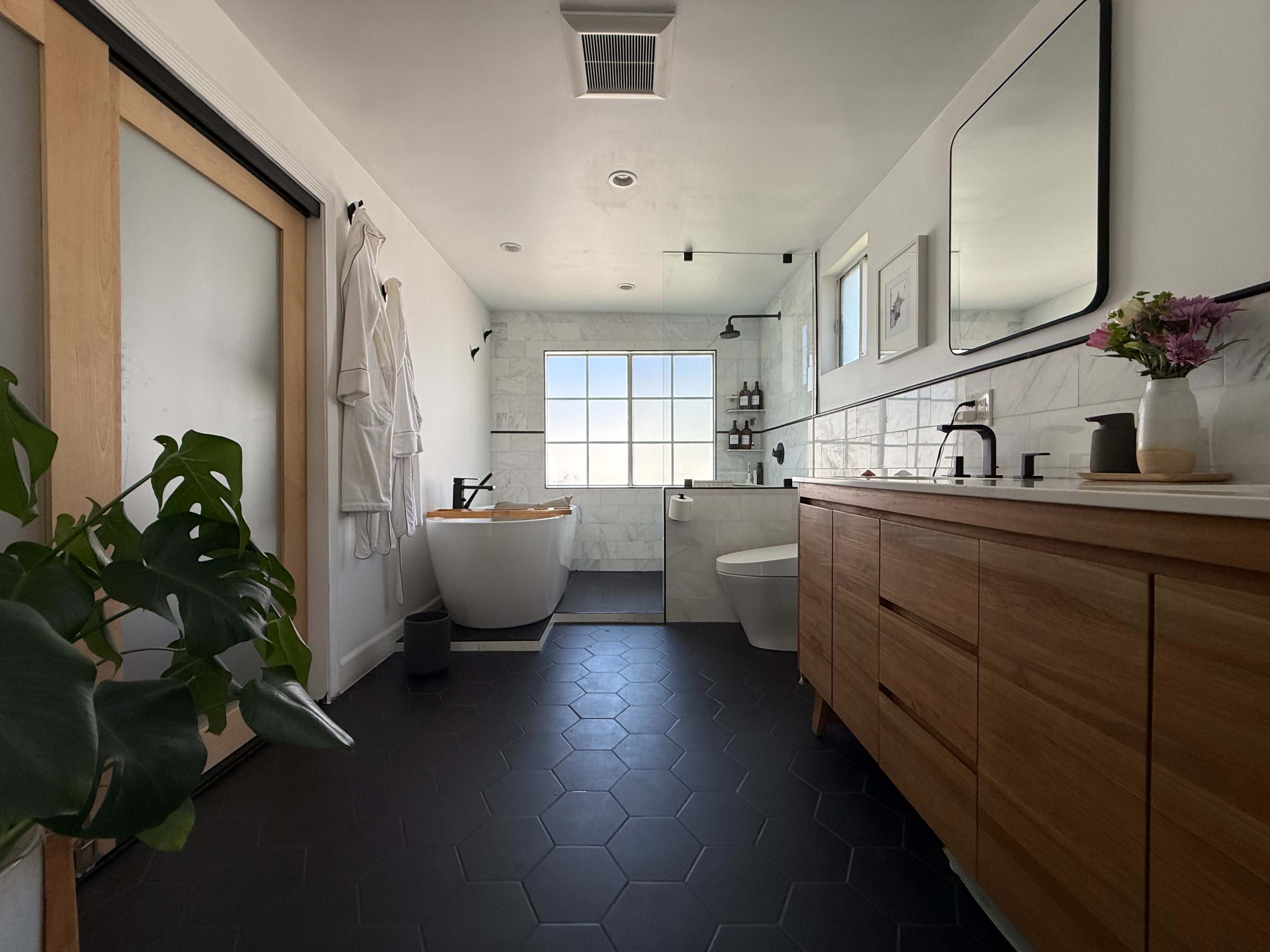 The image features a modern bathroom with a freestanding bathtub, wooden vanity, a glass shower, and a large window providing natural light.