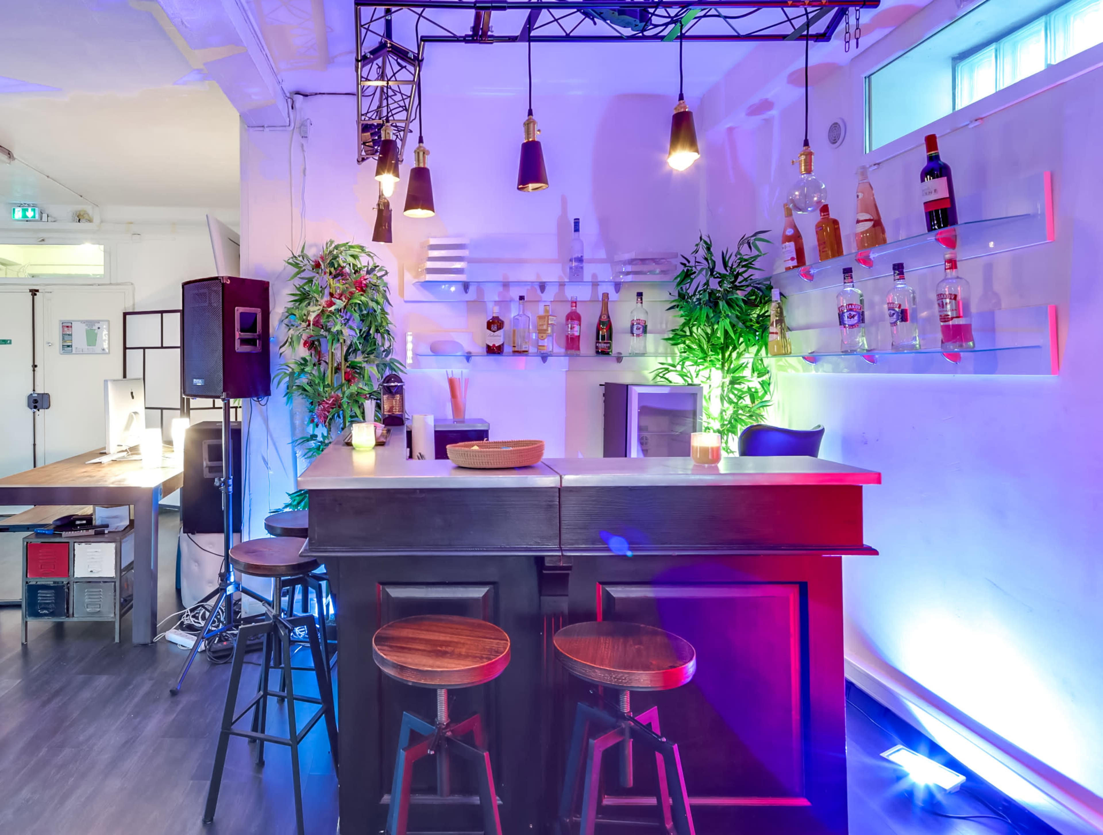 The image shows a modern bar area with a black counter, wooden stools, and illuminated shelves displaying various bottles, all set against a brightly lit backdrop.