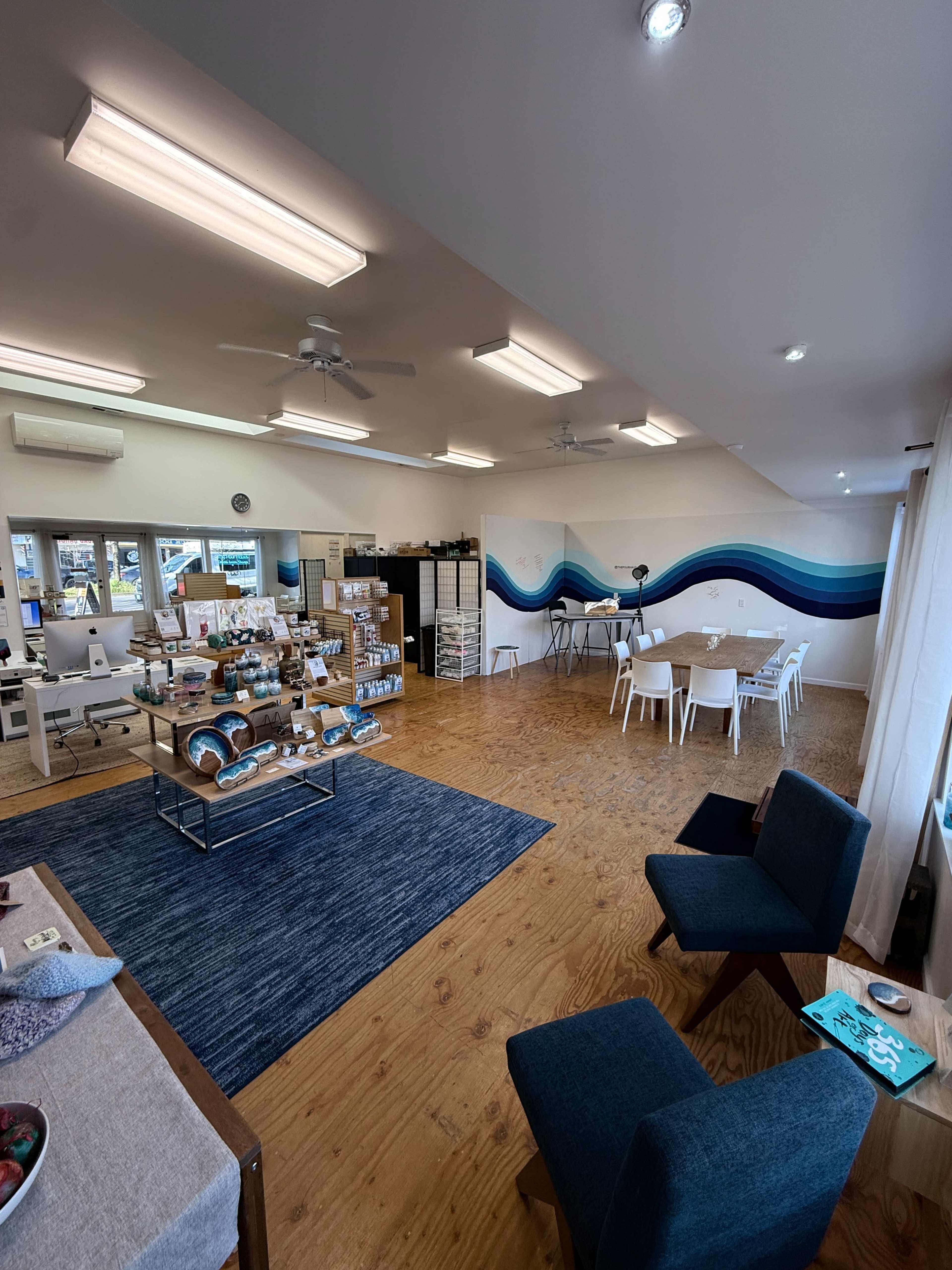 The image shows a bright, spacious room with a wooden floor featuring a seating area, a display table with various products, a dining area with white chairs, and a mural on the wall.