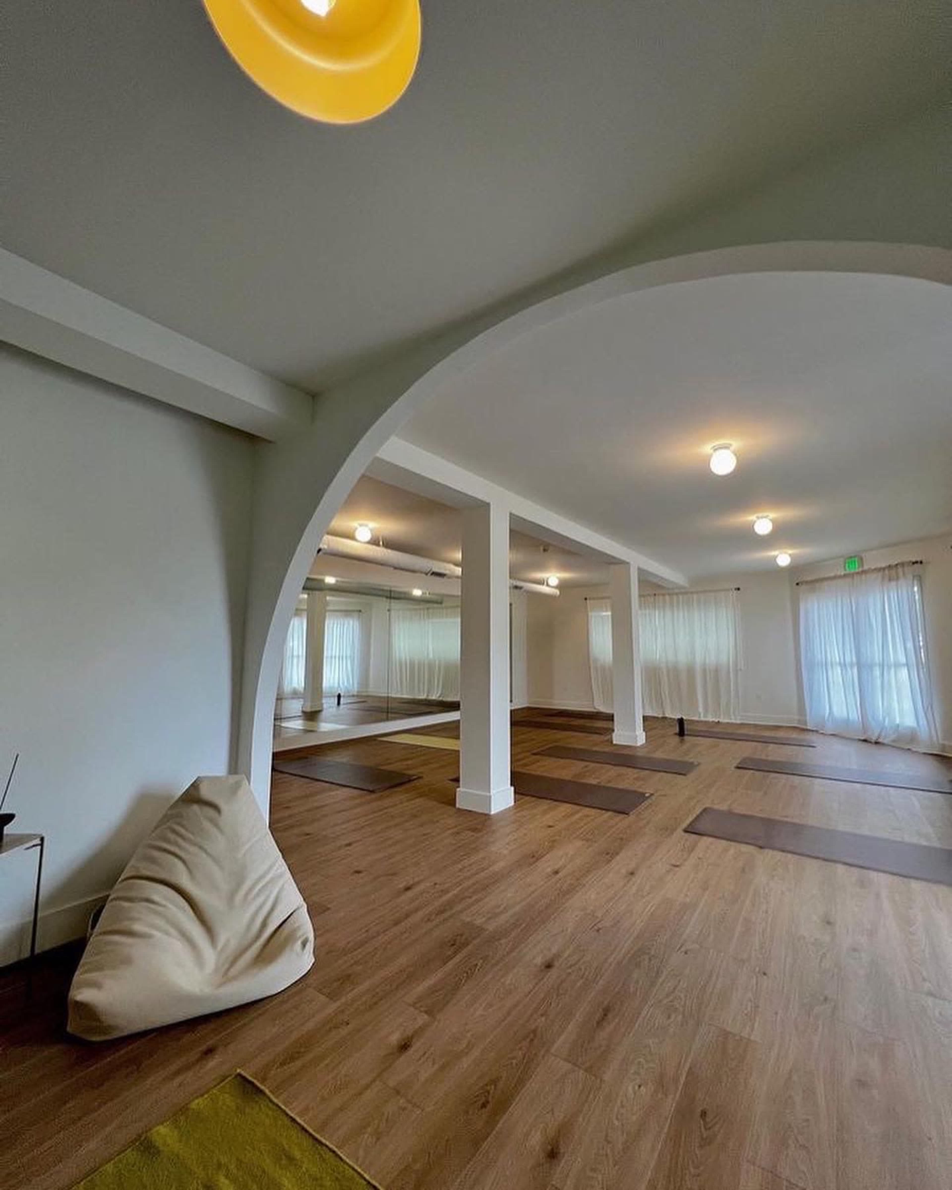 A spacious yoga studio with wooden floors, large windows, and minimal furnishings, including yoga mats and a bean bag chair.