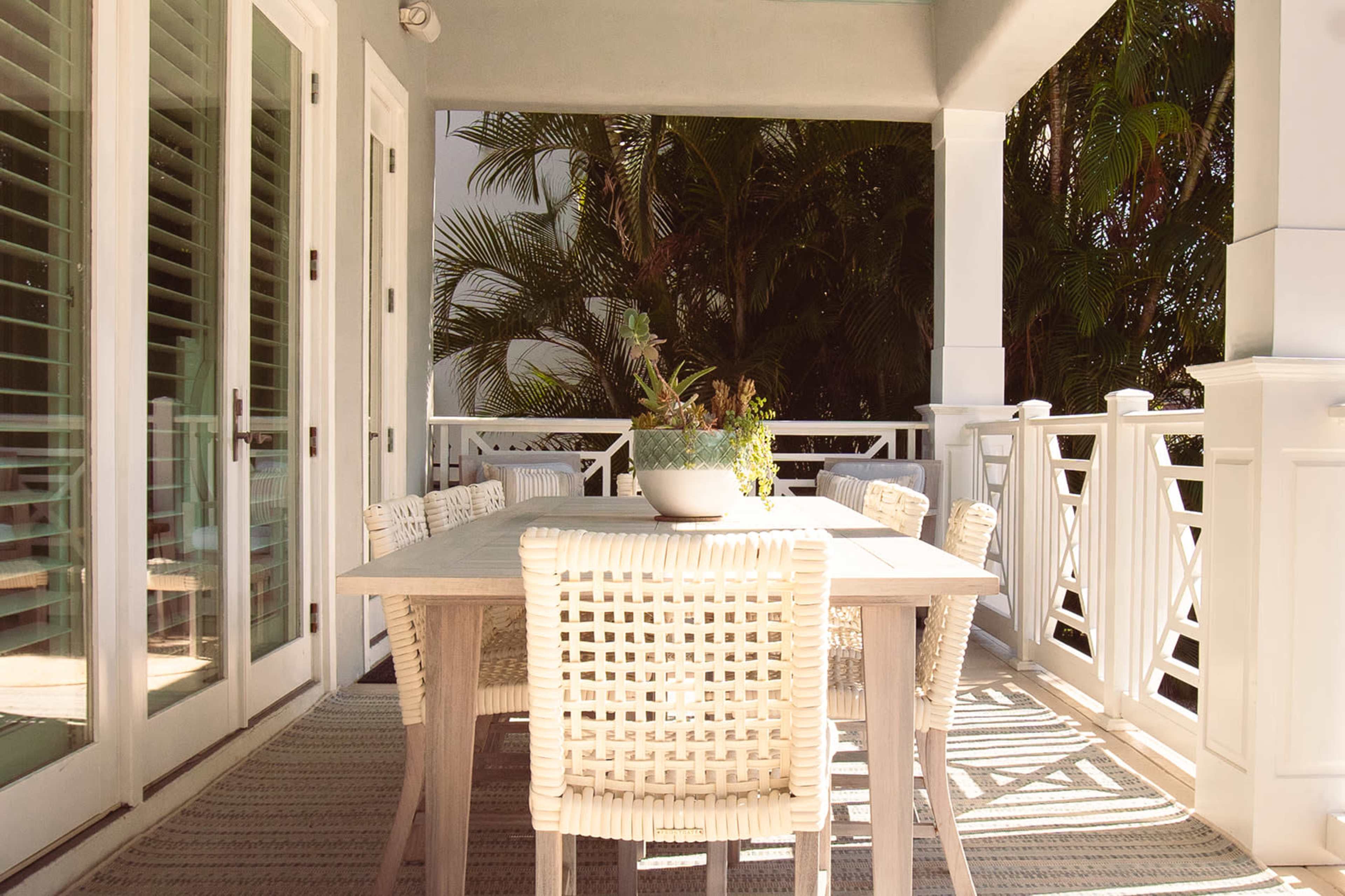 A wooden dining table with woven chairs is set on a porch, surrounded by greenery and sunlight.