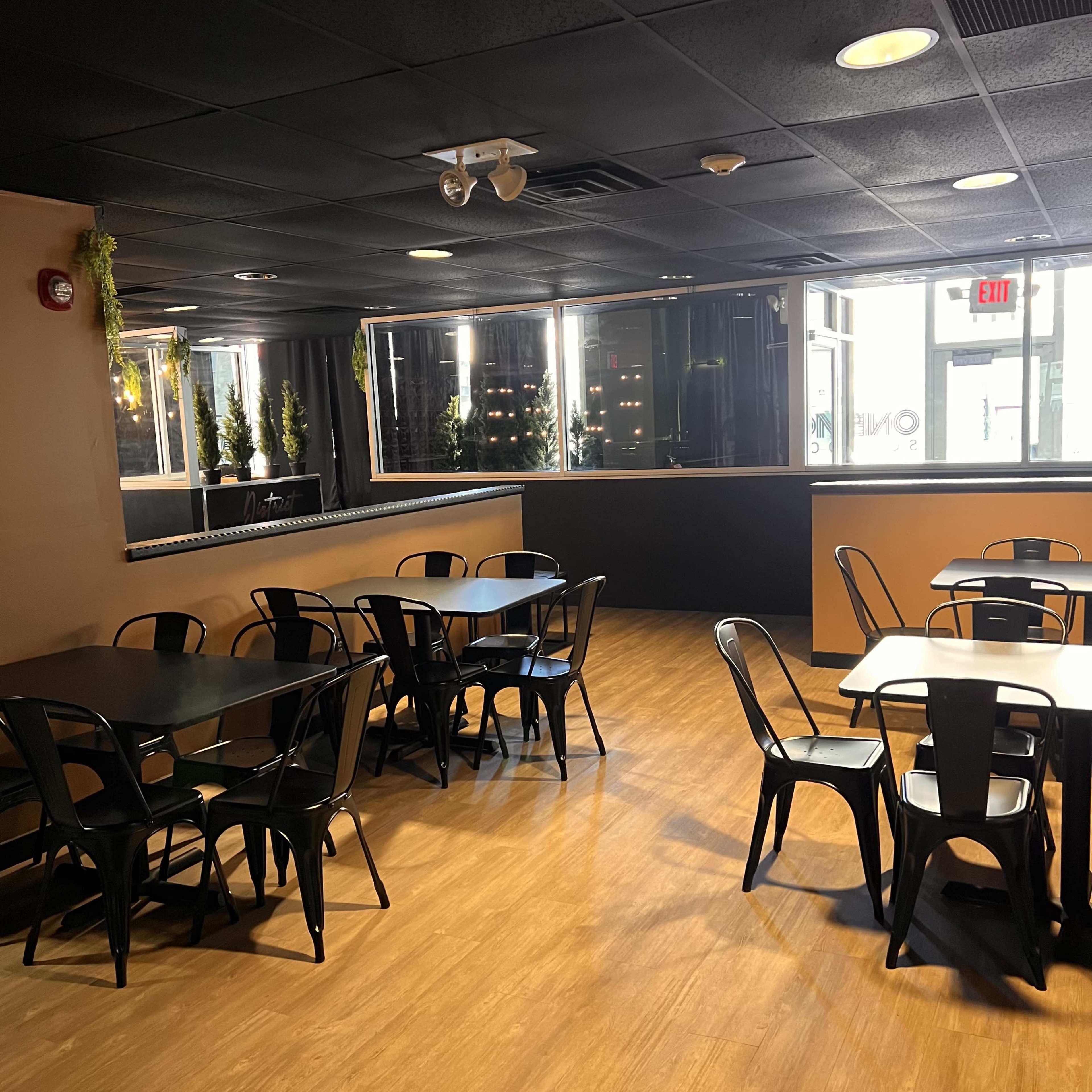 The image shows a modern indoor dining area featuring several black metal tables and chairs, with large windows allowing natural light to enter.