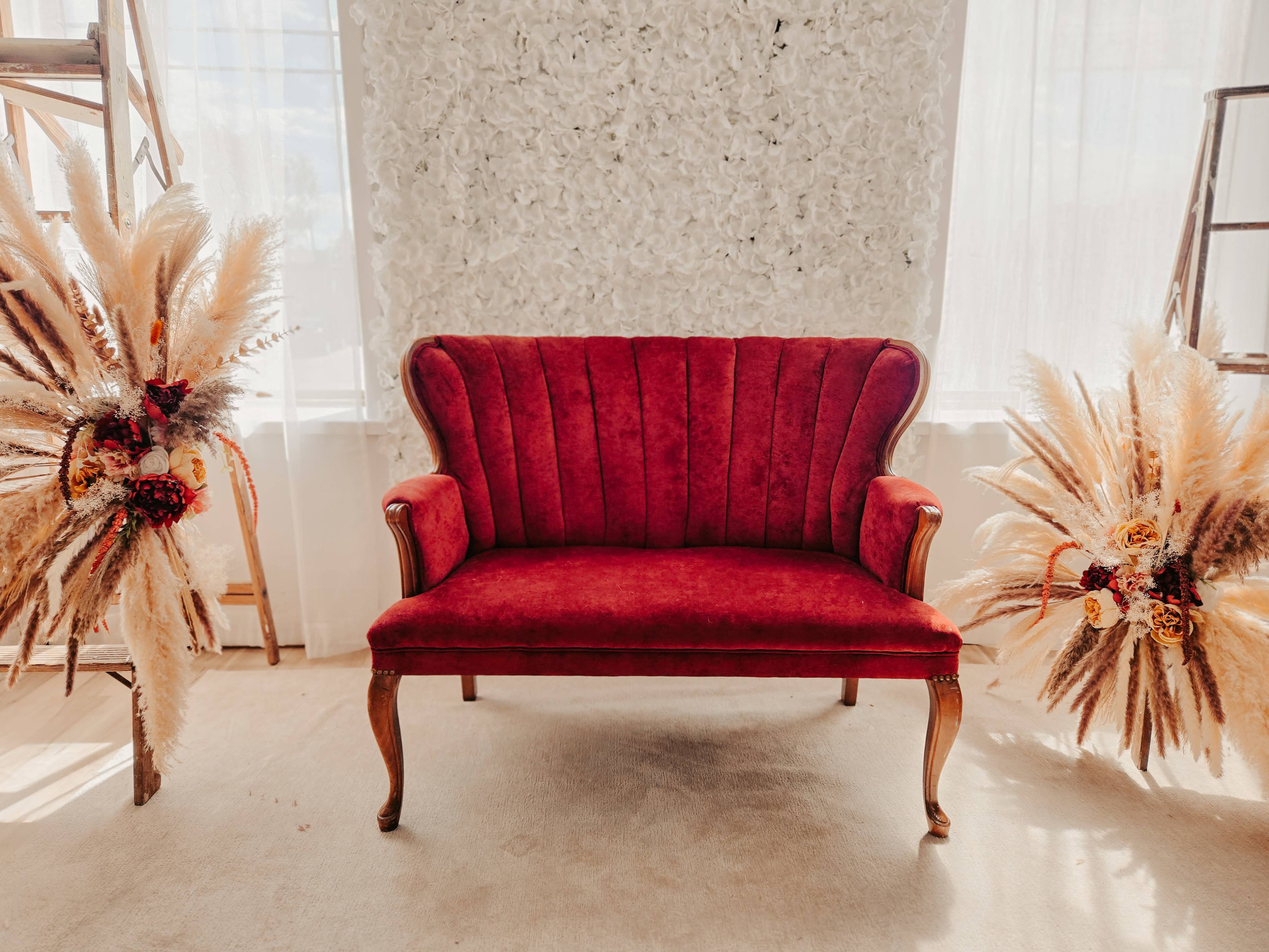 A red velvet loveseat is positioned between floral arrangements made of pampas grass against a light-colored wall.