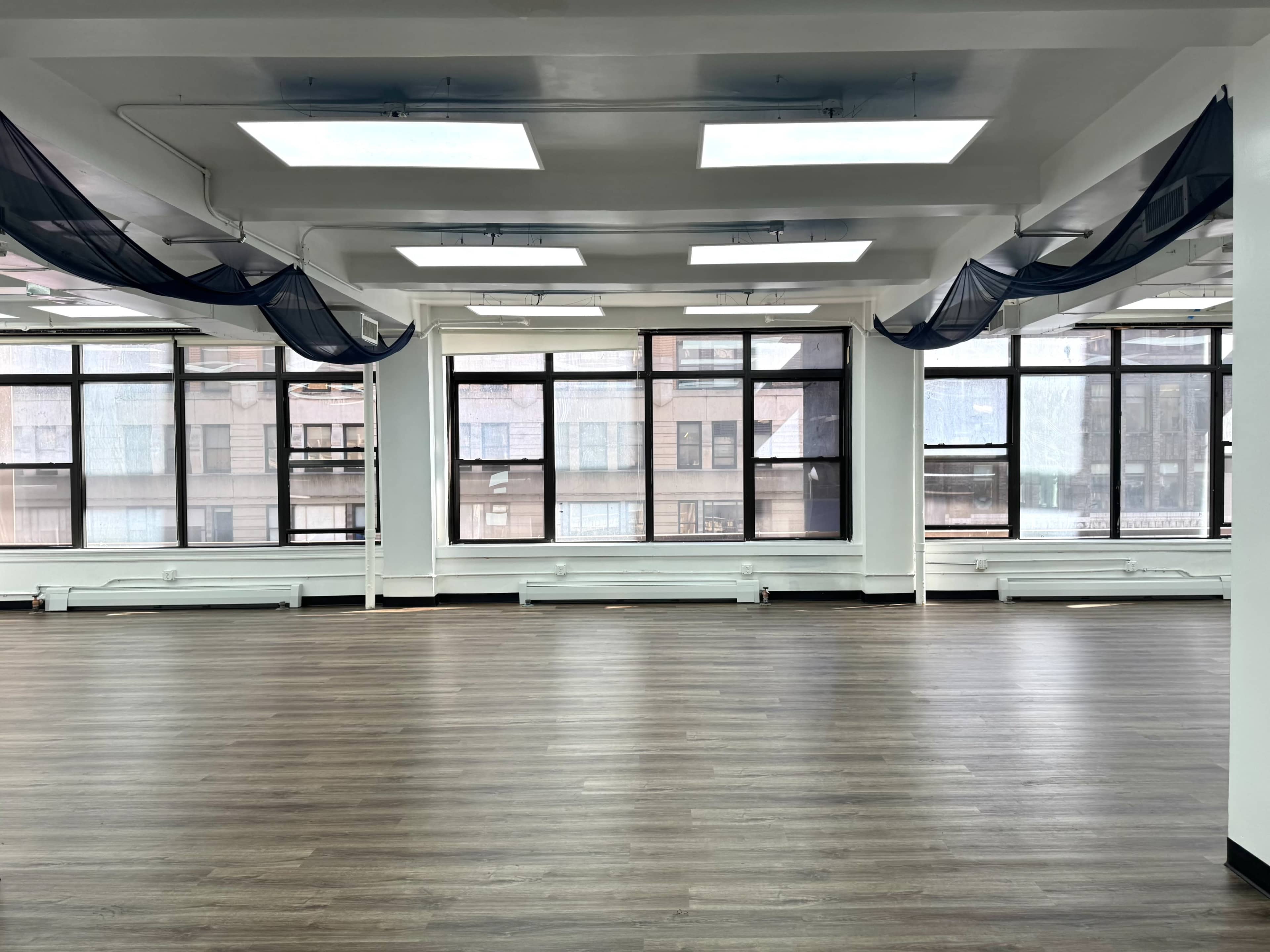 The image shows a spacious room with large windows, modern lighting, and a wooden floor, featuring sheer curtains draped from the ceiling.