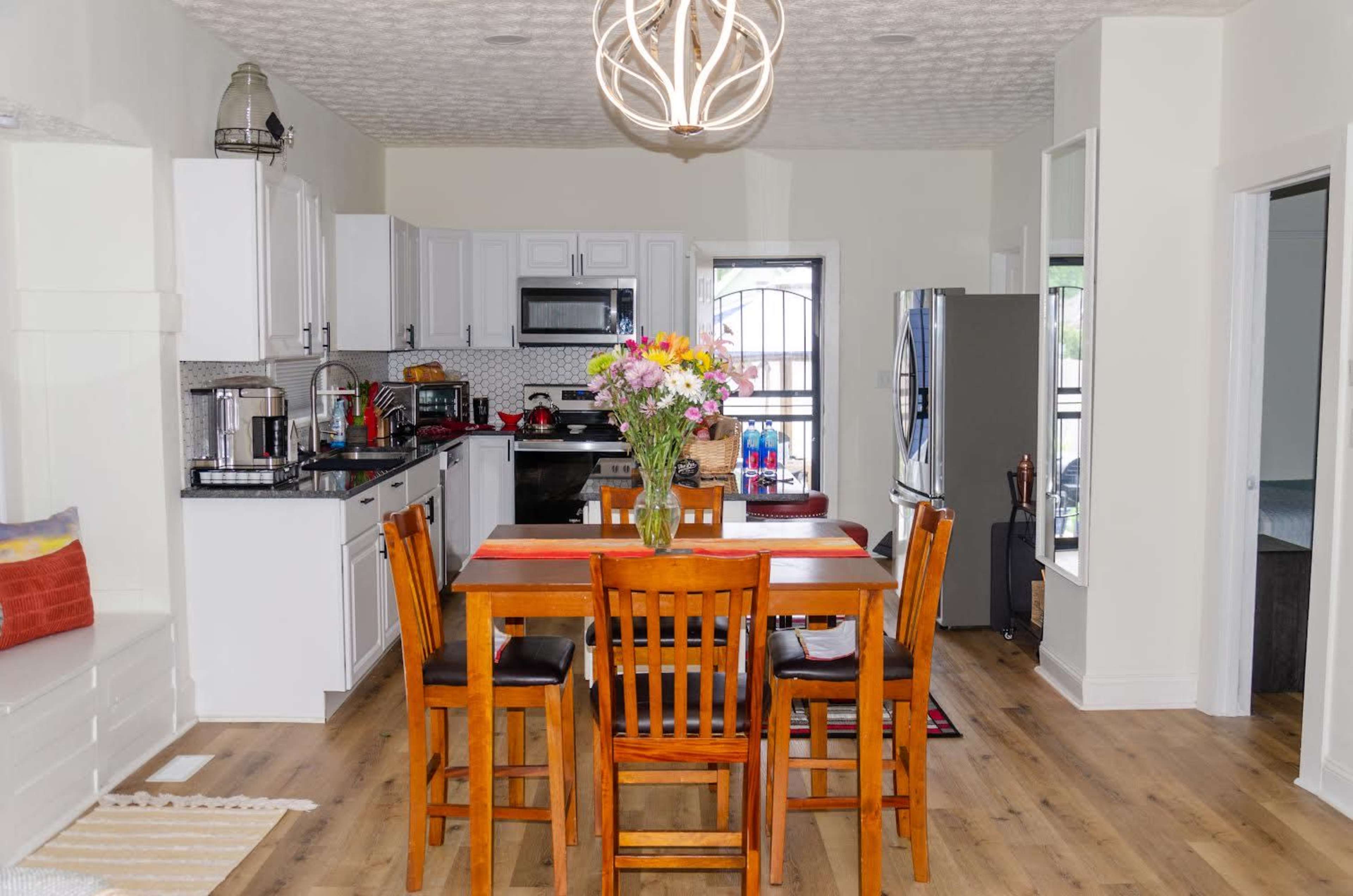 A modern kitchen features white cabinets, stainless steel appliances, a wooden dining table with four chairs, and a vase of flowers in the center.