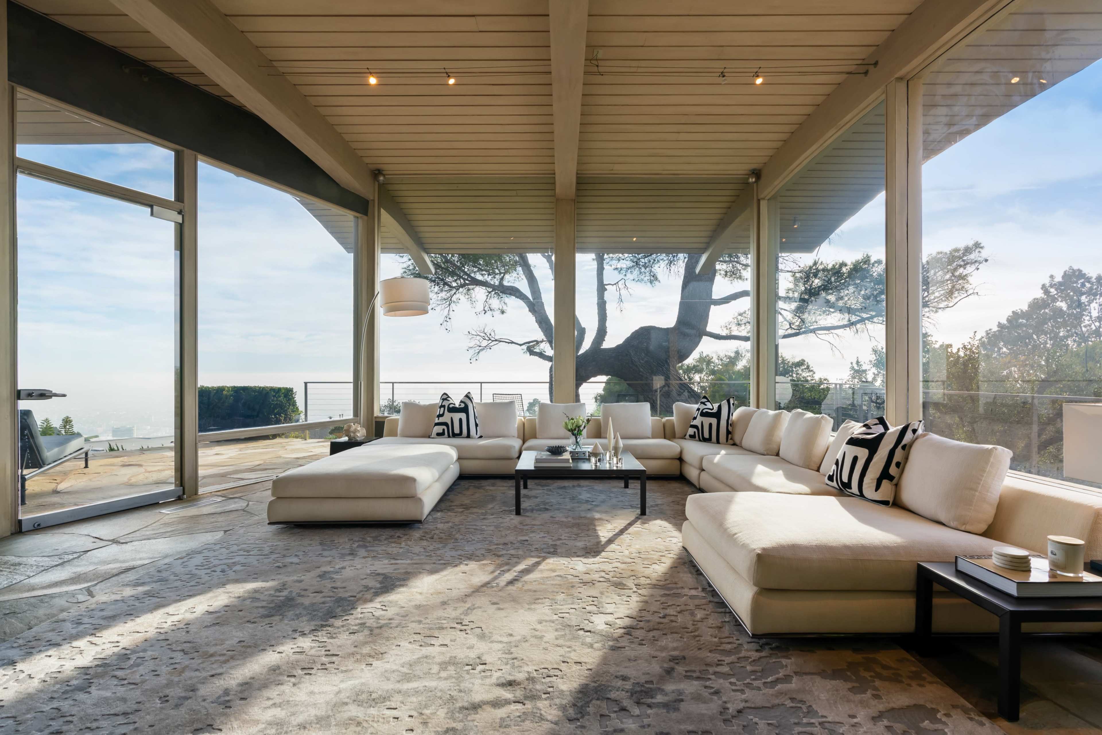 A spacious, modern living room features a large sectional sofa, a coffee table, and expansive glass walls that overlook a scenic outdoor view.