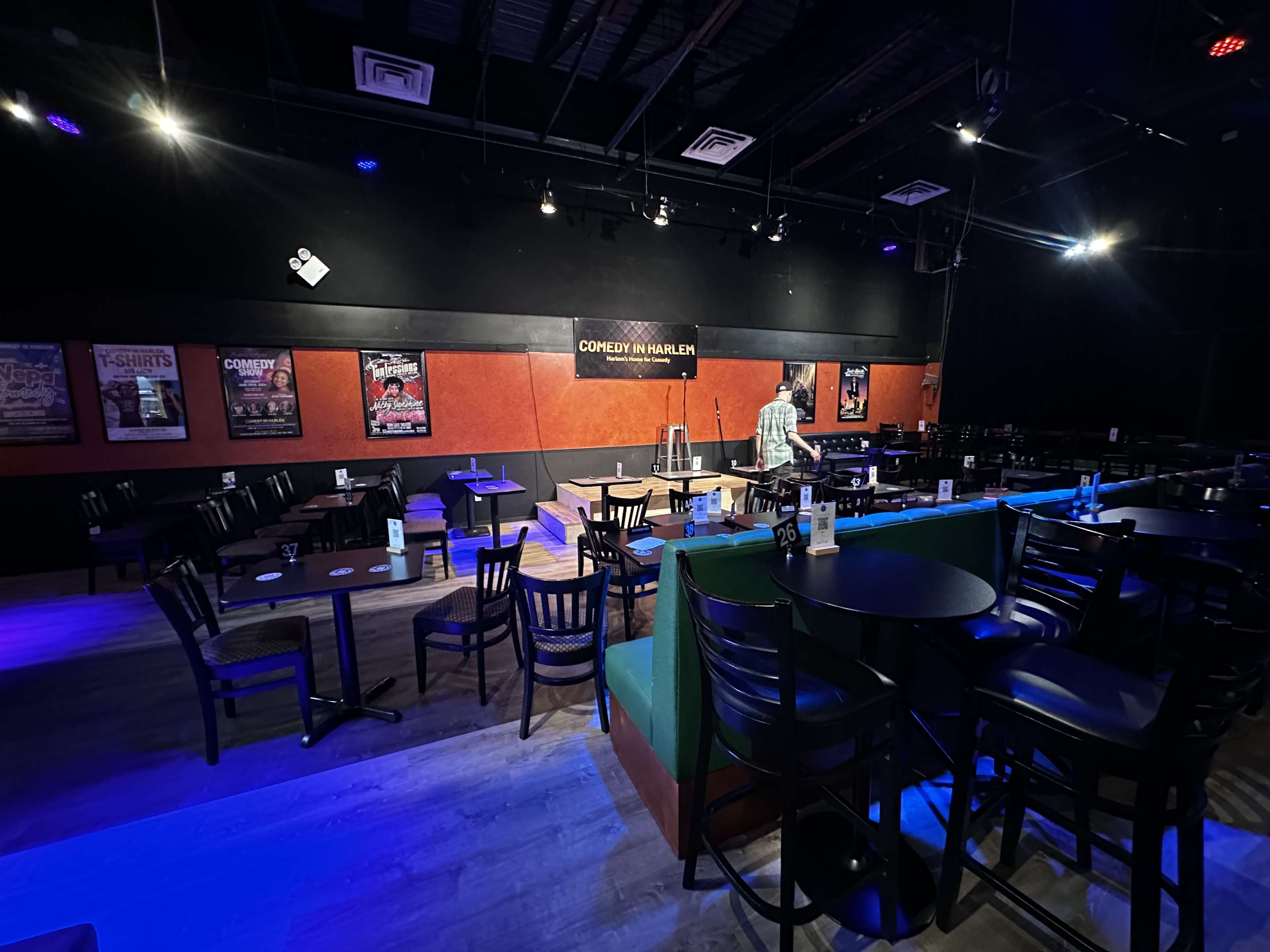 The image shows a dimly lit comedy club with empty tables and chairs arranged in front of a small stage, where a person is walking towards a microphone.