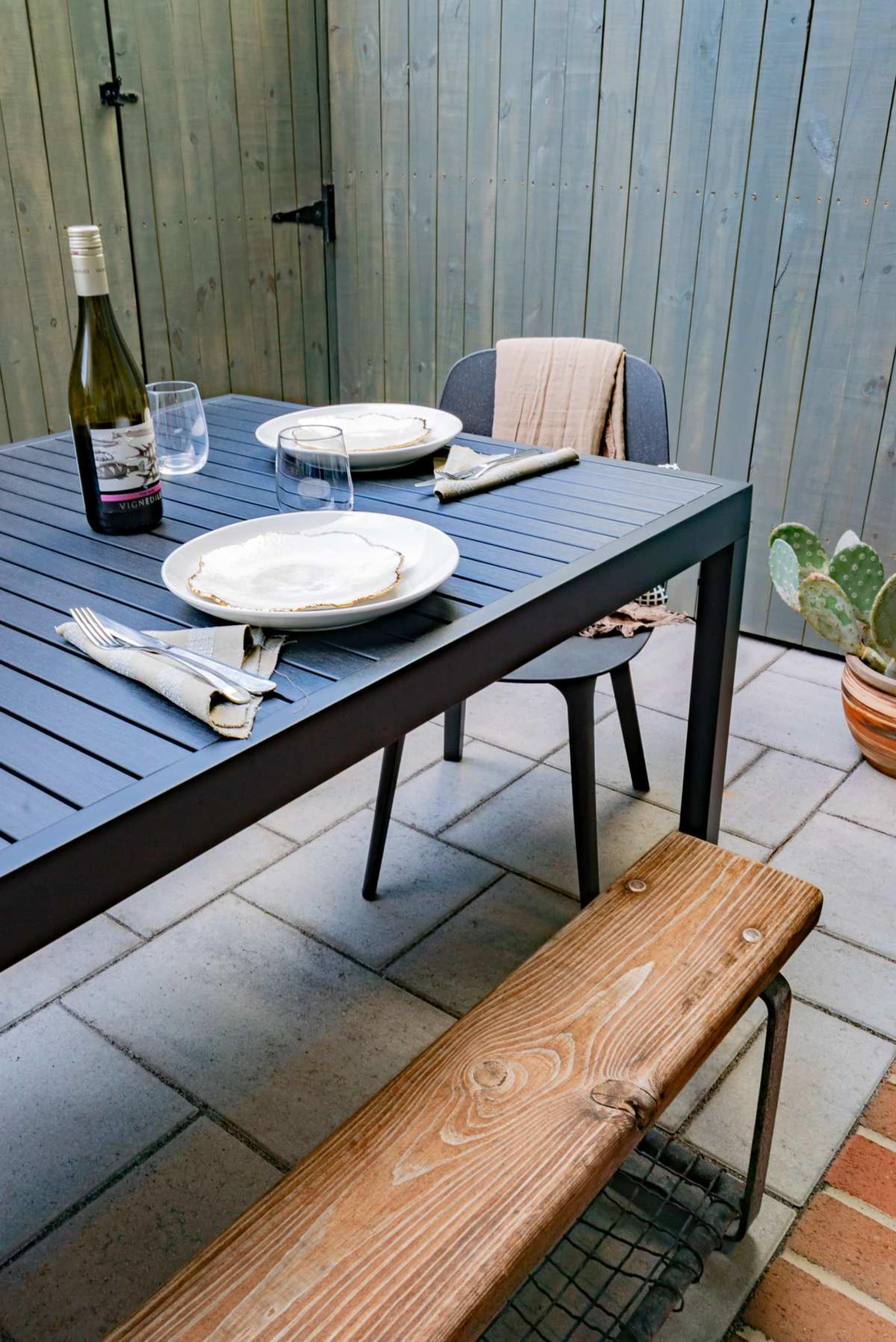 A gray outdoor dining table set with plates, glasses, and a bottle of wine is positioned on a tiled patio surrounded by wooden fencing and a potted cactus.